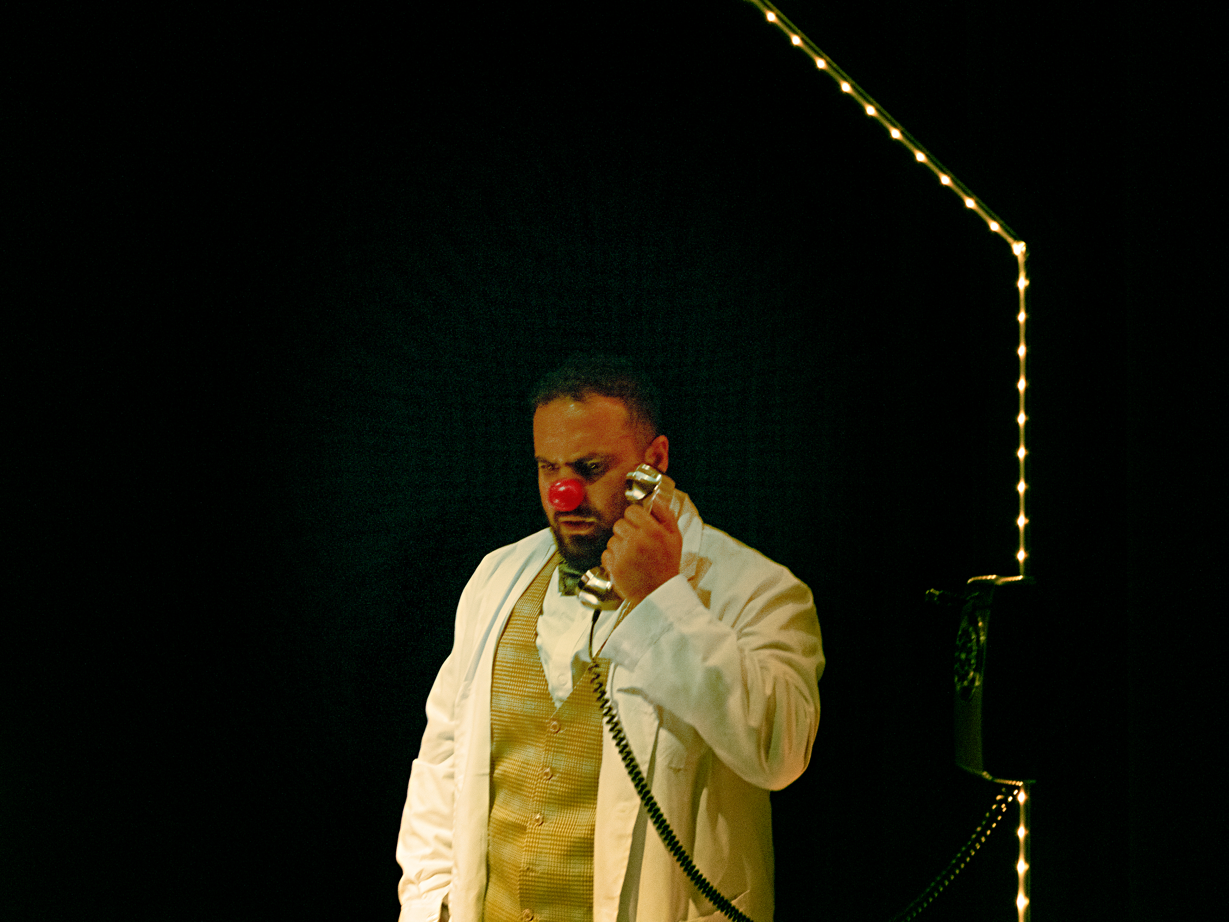 A man with a clown nose, makeup, and a white shirt holding a telephone, standing in a dimly lit room with string lights hanging in the background.