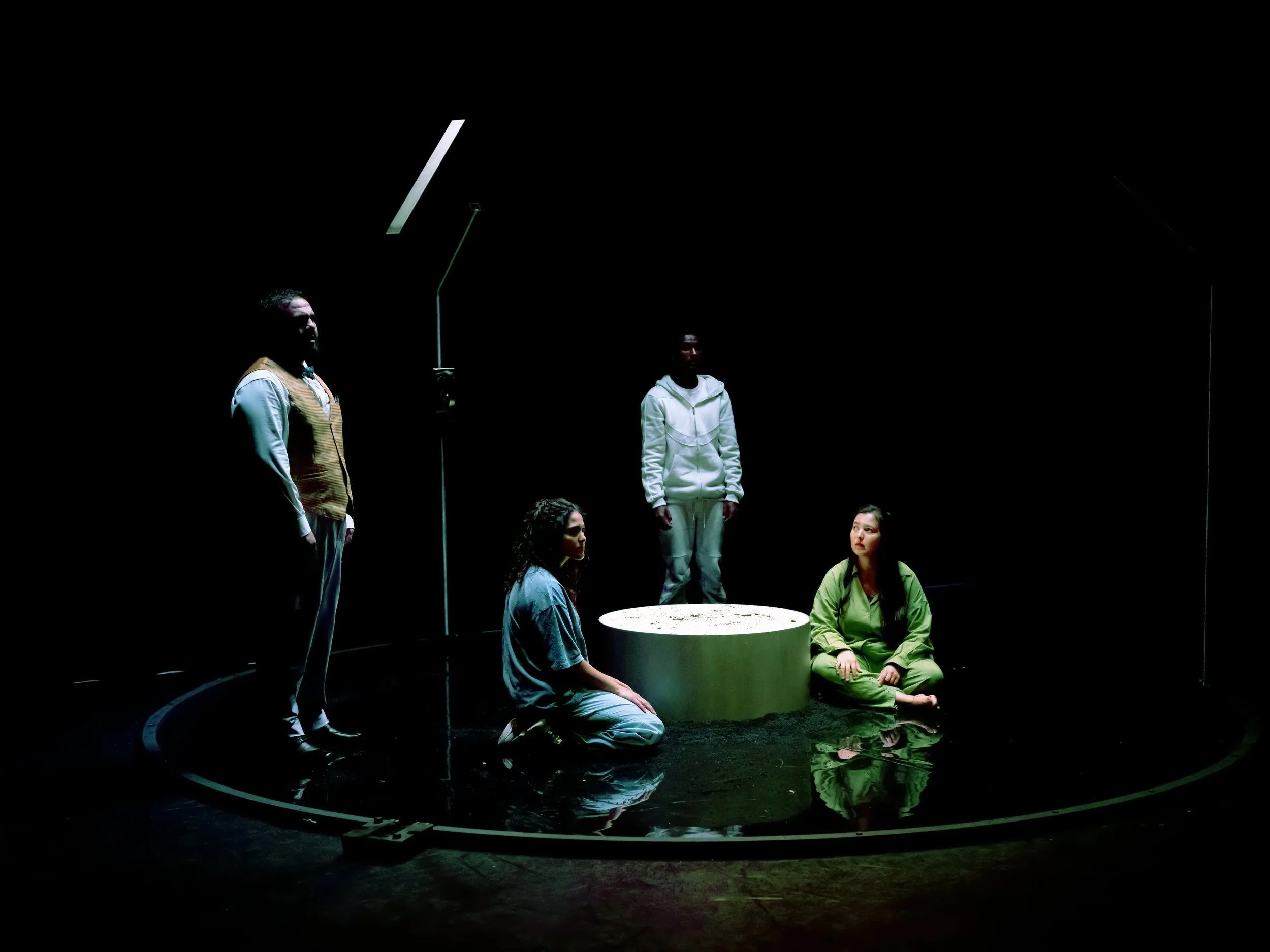 Scene from a theatrical performance showing four actors in a dark setting, one standing, three seated on the floor, with a circular water feature in the foreground and a white circular object in the center.