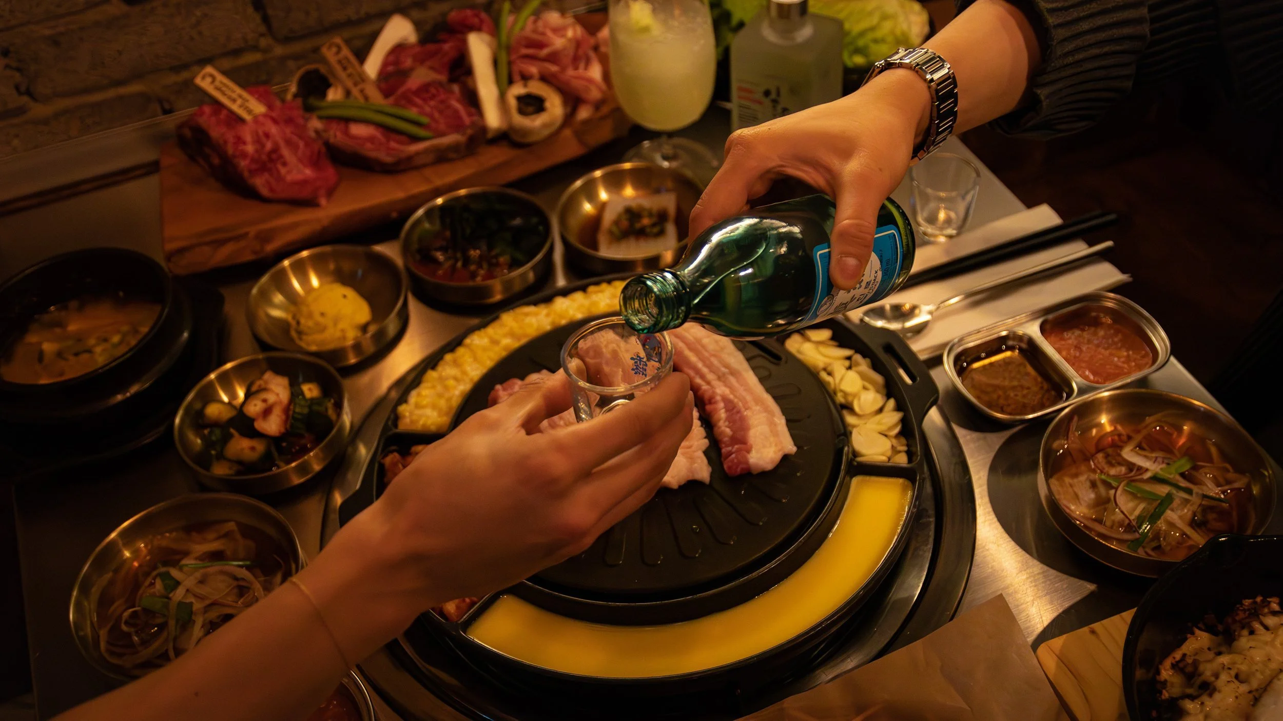 Person pouring a drink into a shot glass at a table filled with various dishes, including raw meat, vegetables, and sauces, in a dimly lit restaurant.