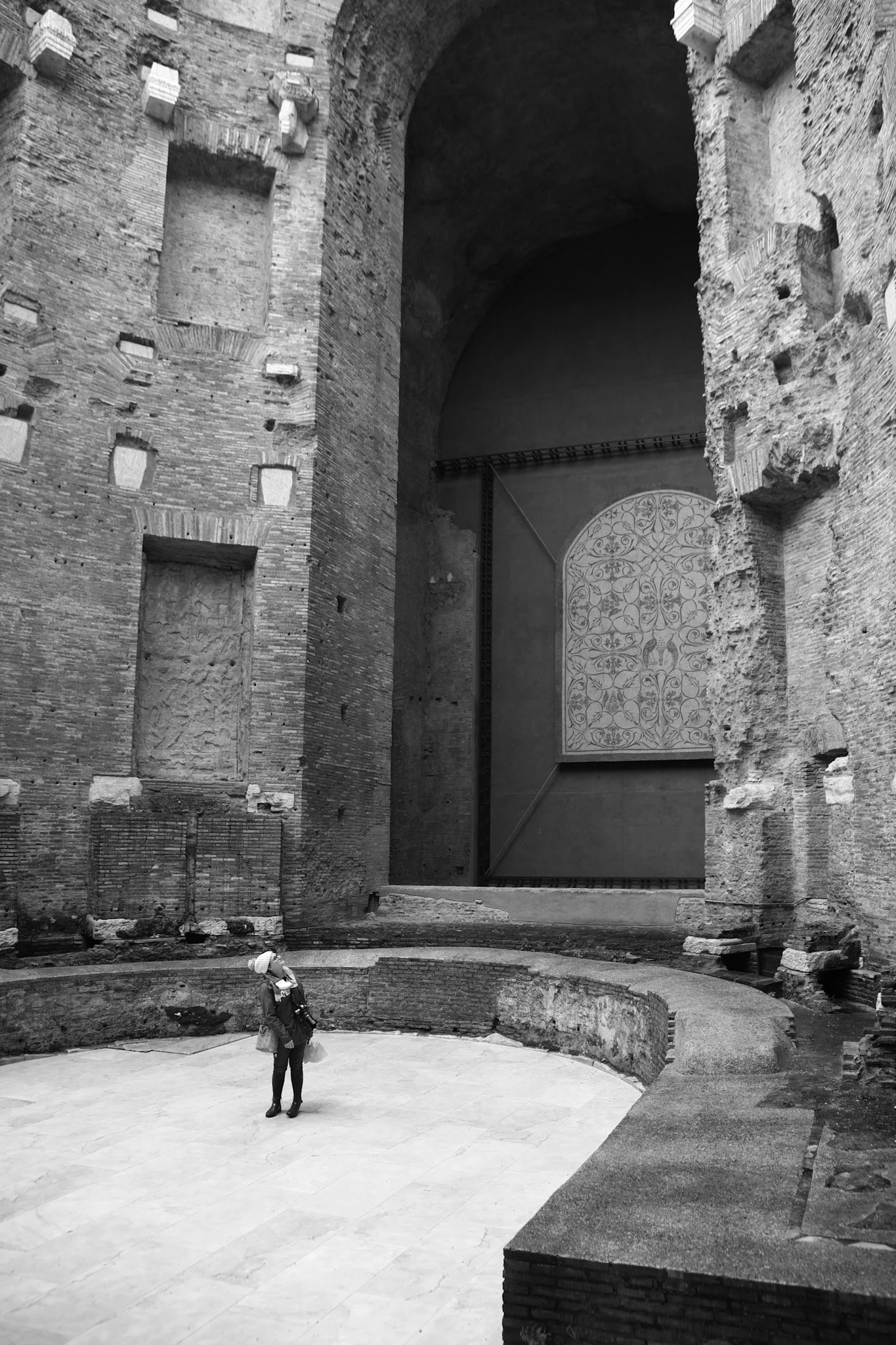 Ancient ruins with brick and stone walls, a large arch, and a decorative wall panel. A person in a white hat and dark clothing stands in the foreground. The environment appears to be an archaeological site or historical structure.