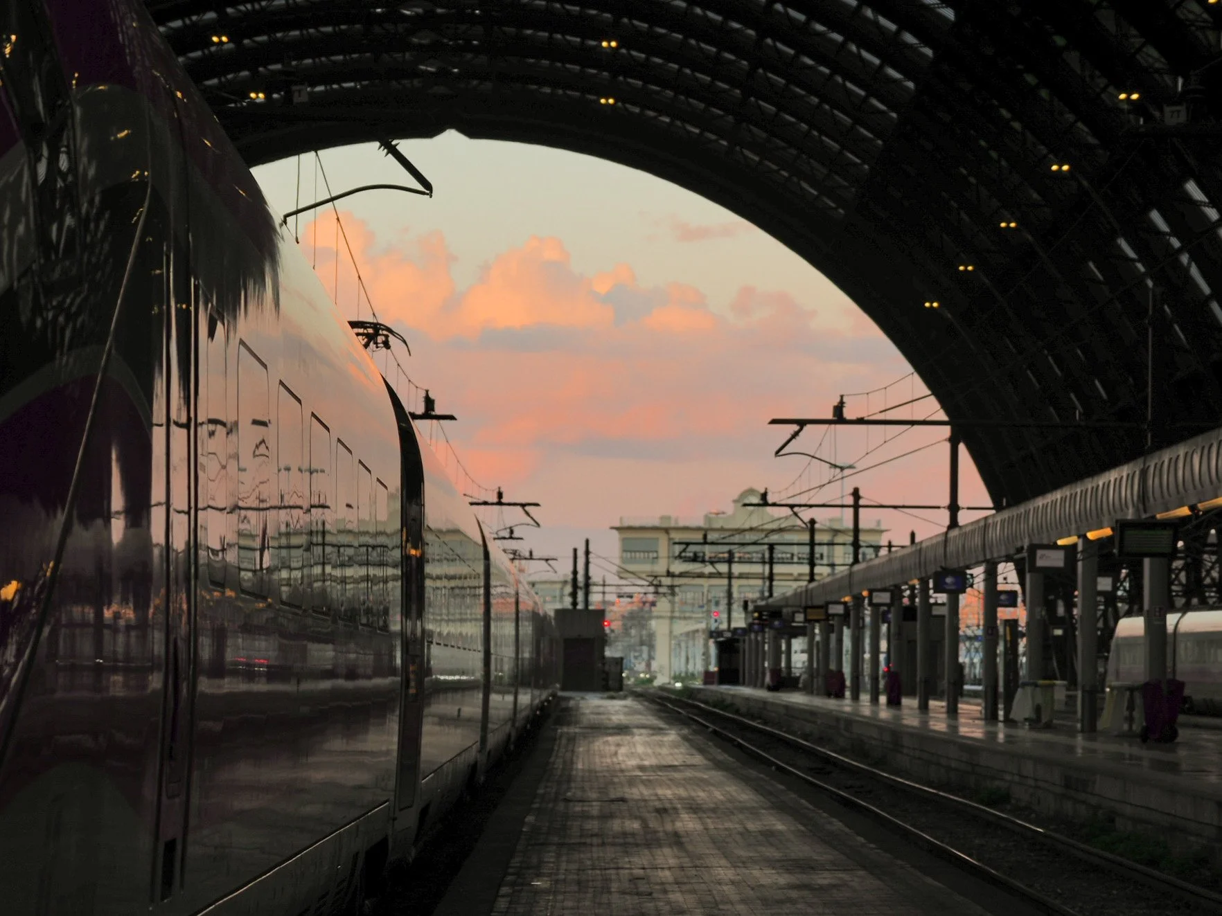 A train station at sunset with an exposed overhead arch, train platforms, and electric wires, reflecting the sky with pink and orange clouds.