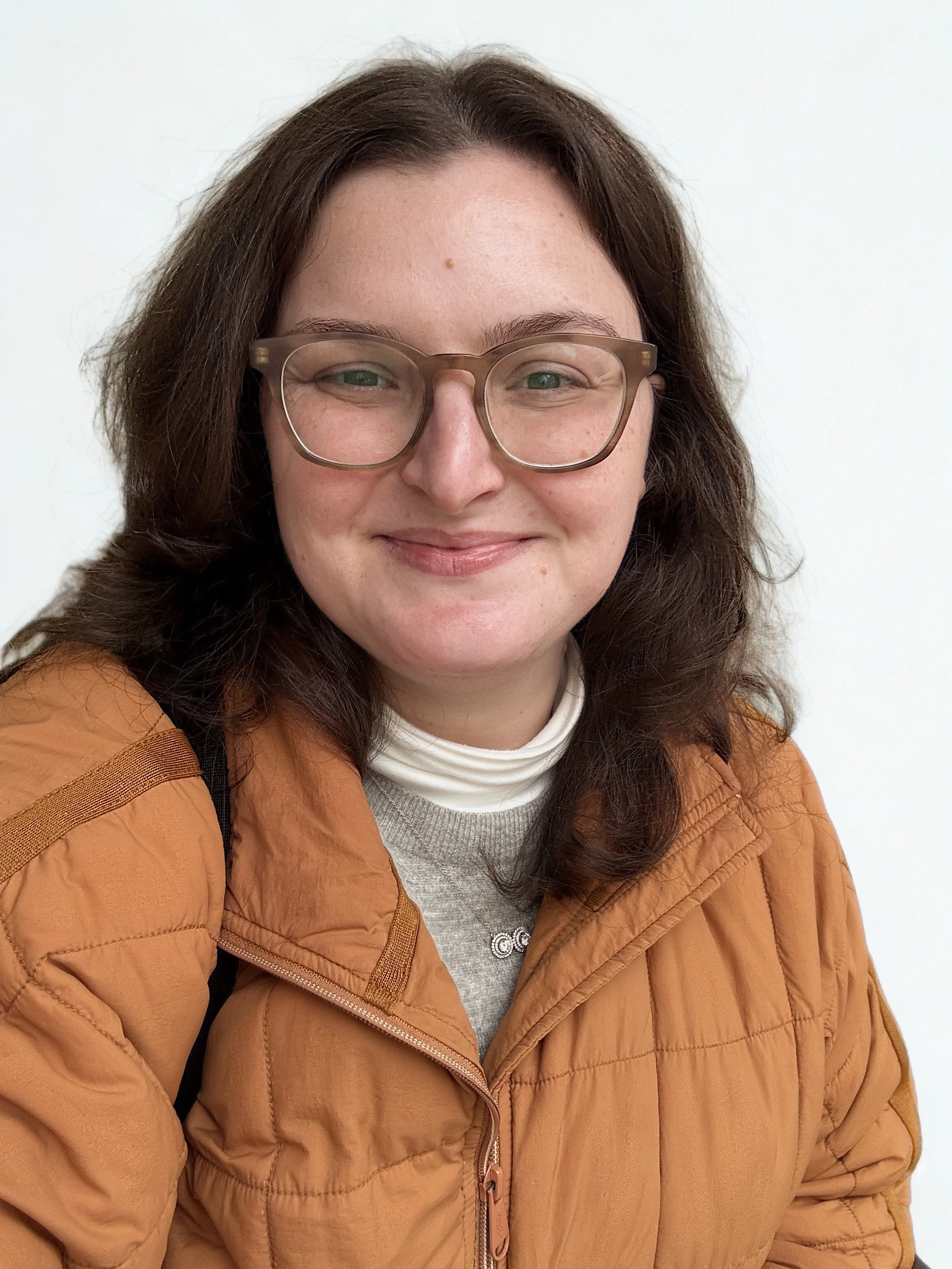 A woman with brown hair, wearing glasses, a white turtleneck, and a tan quilted jacket, smiling at the camera against a plain white background.