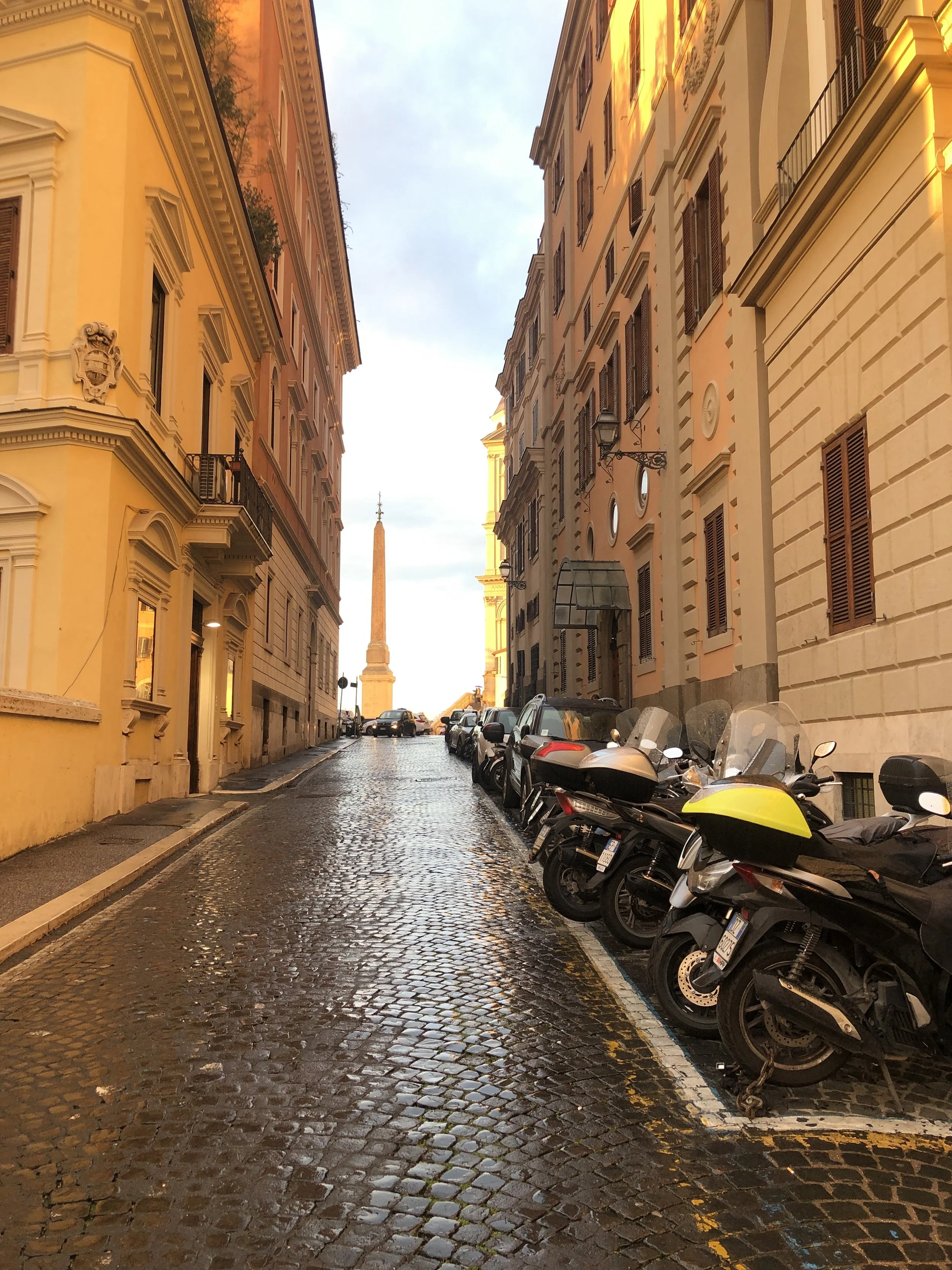 A cobblestone street lined with parked motorcycles on the right side, yellow and orange historic buildings on the left and right, and a view of a tall obelisk monument in the distance with a cloudy sky above at dusk.