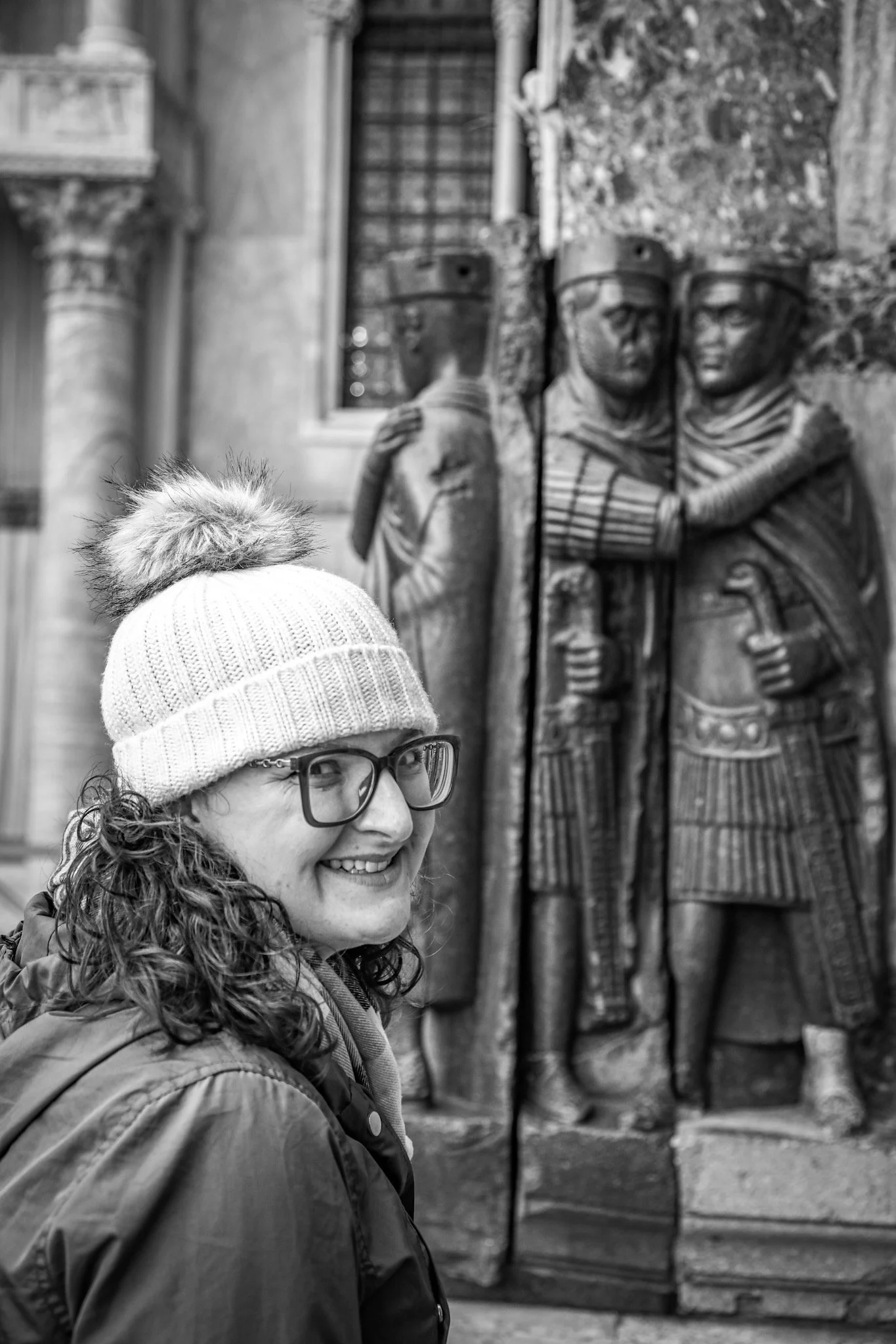 A woman smiling, wearing glasses, a knit hat with a pom-pom, a jacket, and a scarf, standing outdoors near a historic stone wall with carved sculptures of tetrarchs.