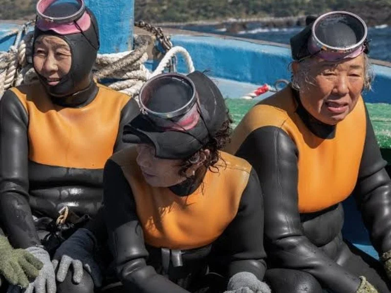 Three women wearing wetsuits and snorkeling gear on a boat near the water.
