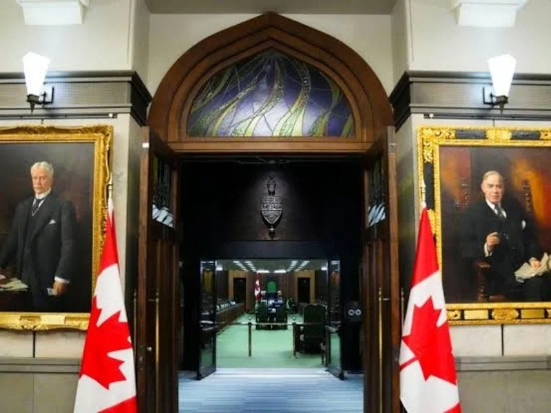 View into a government chamber, with portraits of officials on either side, Canadian flags, and seating for members.