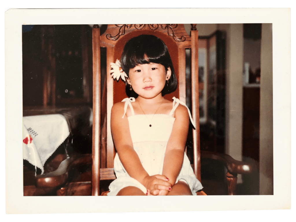 A young Hannah Sung sitting on a wooden chair with a high backrest, wearing a white dress, in a room with wooden furniture and a white tablecloth in the background.