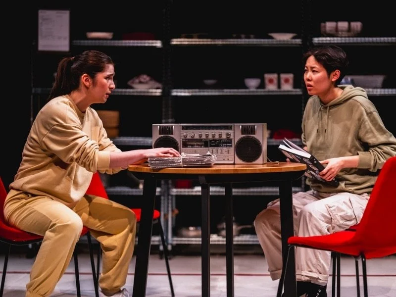 Two women sitting at a table in a room with shelves of dishes, engaged in a conversation, with a radio/tape player on the table.