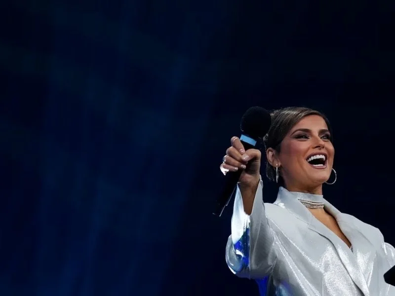 Nelly Furtado smiling and holding a microphone on stage, wearing a shiny silver jacket and hoop earrings.