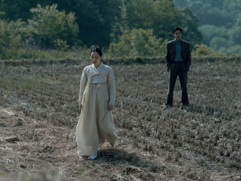 A woman walking through a field while a man in a suit stands in the background.