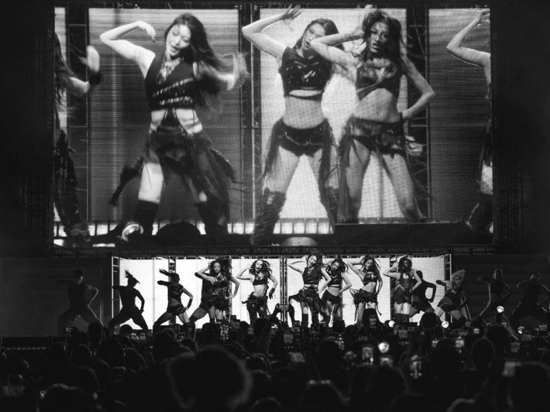 Group of dancers performing on stage with large screens behind them showing close-up images of their dance moves, audience watching in the dark.