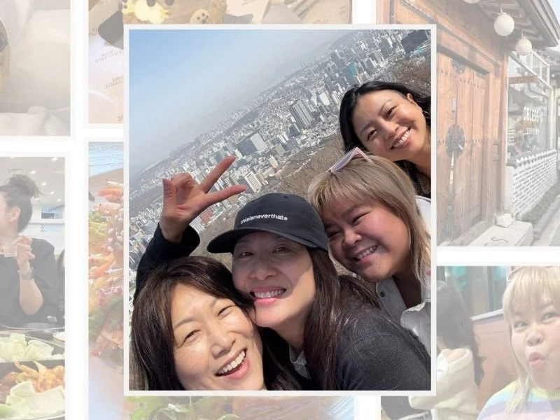 Four women smiling for a selfie outdoors with cityscape in the background.