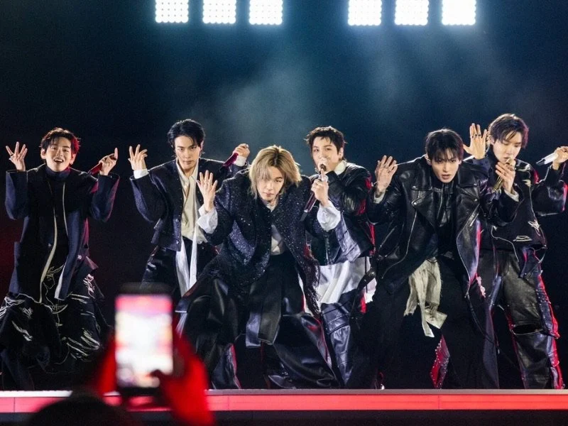 Group of seven performers on stage dressed in dark, stylish clothing, mid-dance move during a concert or performance with bright stage lights overhead.
