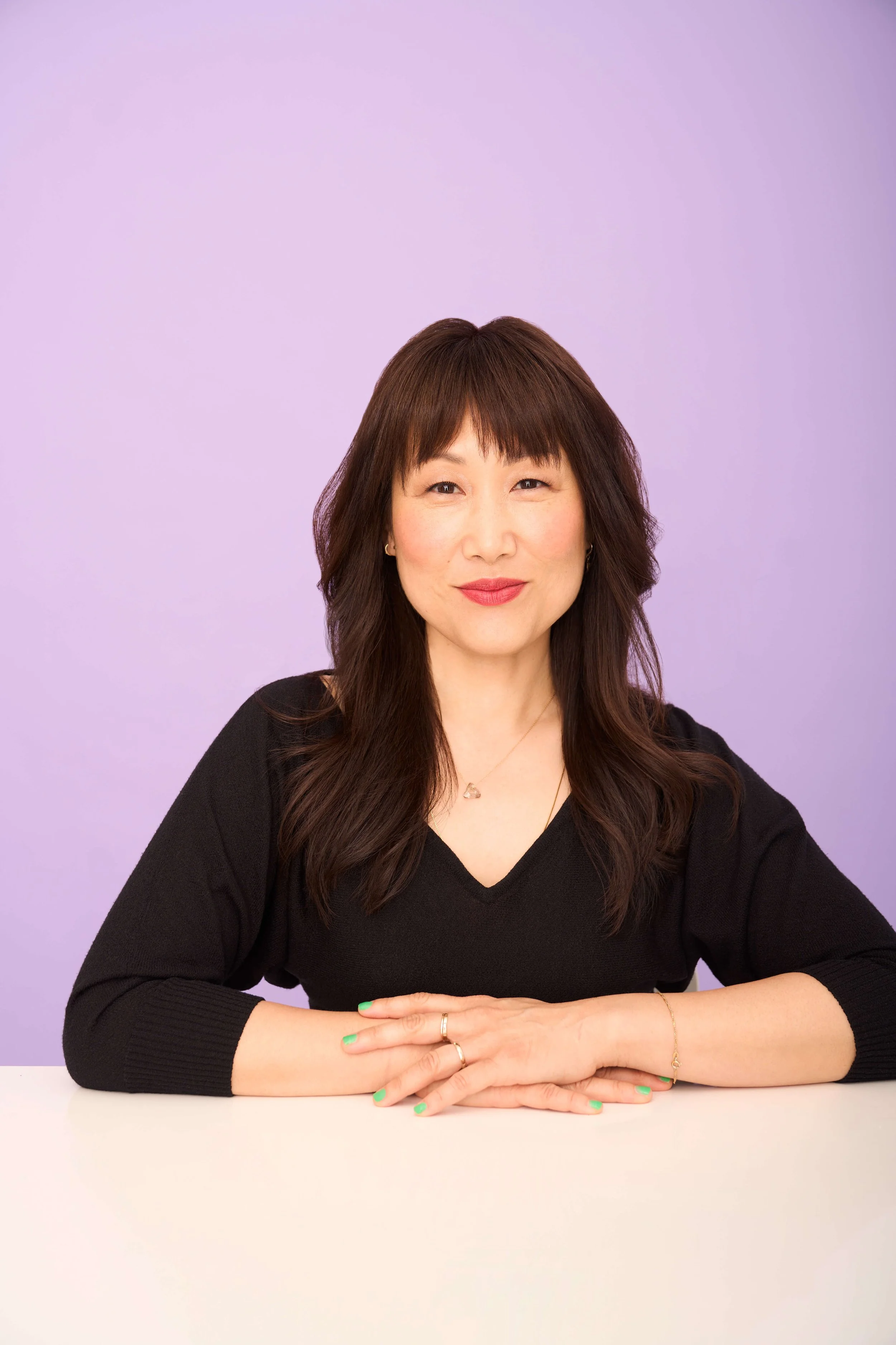 Hannah Sung sitting at a white table with her hands folded and resting on it, against a lavender background.