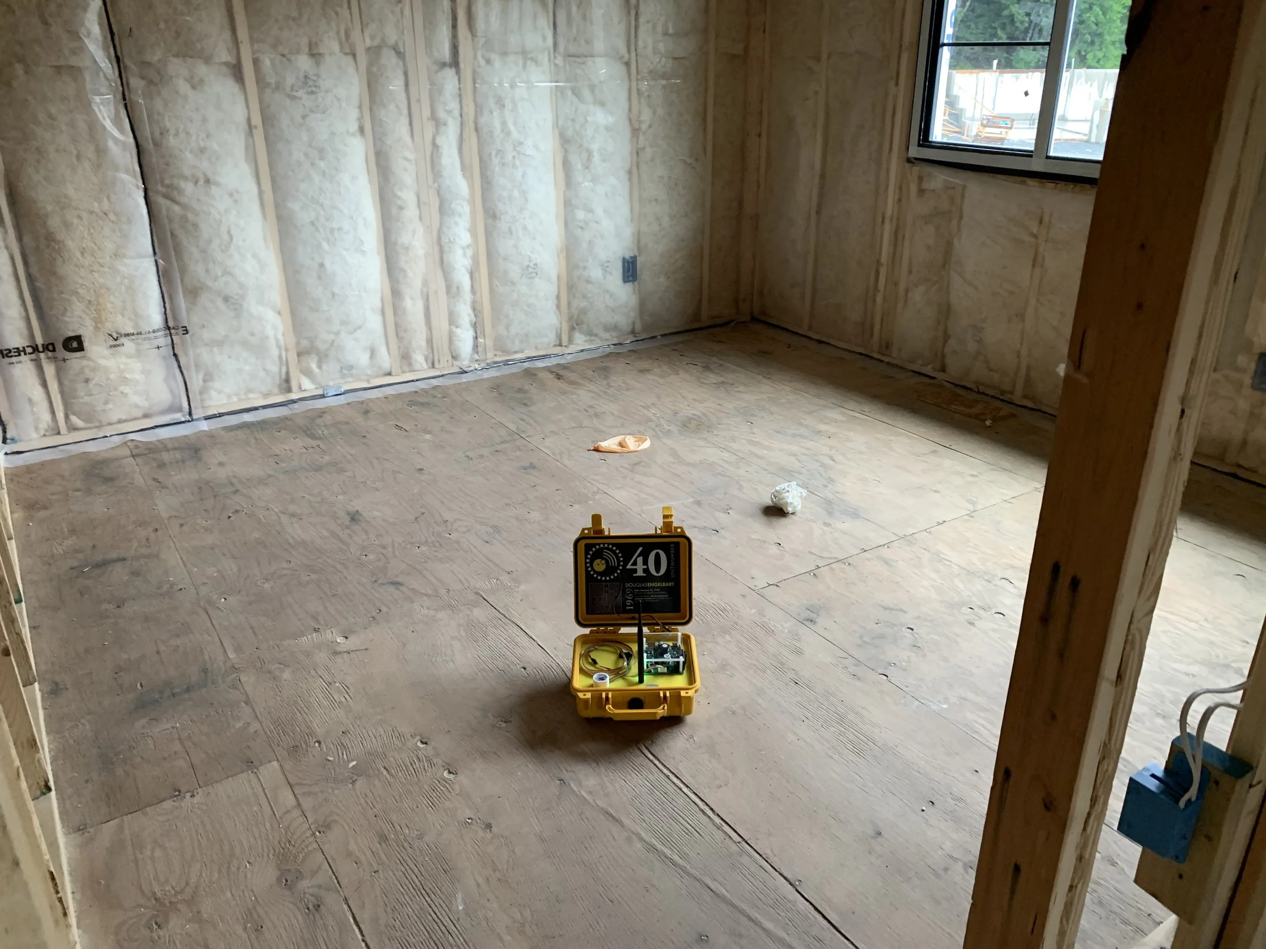 An unfinished room with exposed insulation on the walls, a window to the outside, and a yellow Canary sensor on the floor. This room is about to be disinfected and treated for mold and smoke damage.