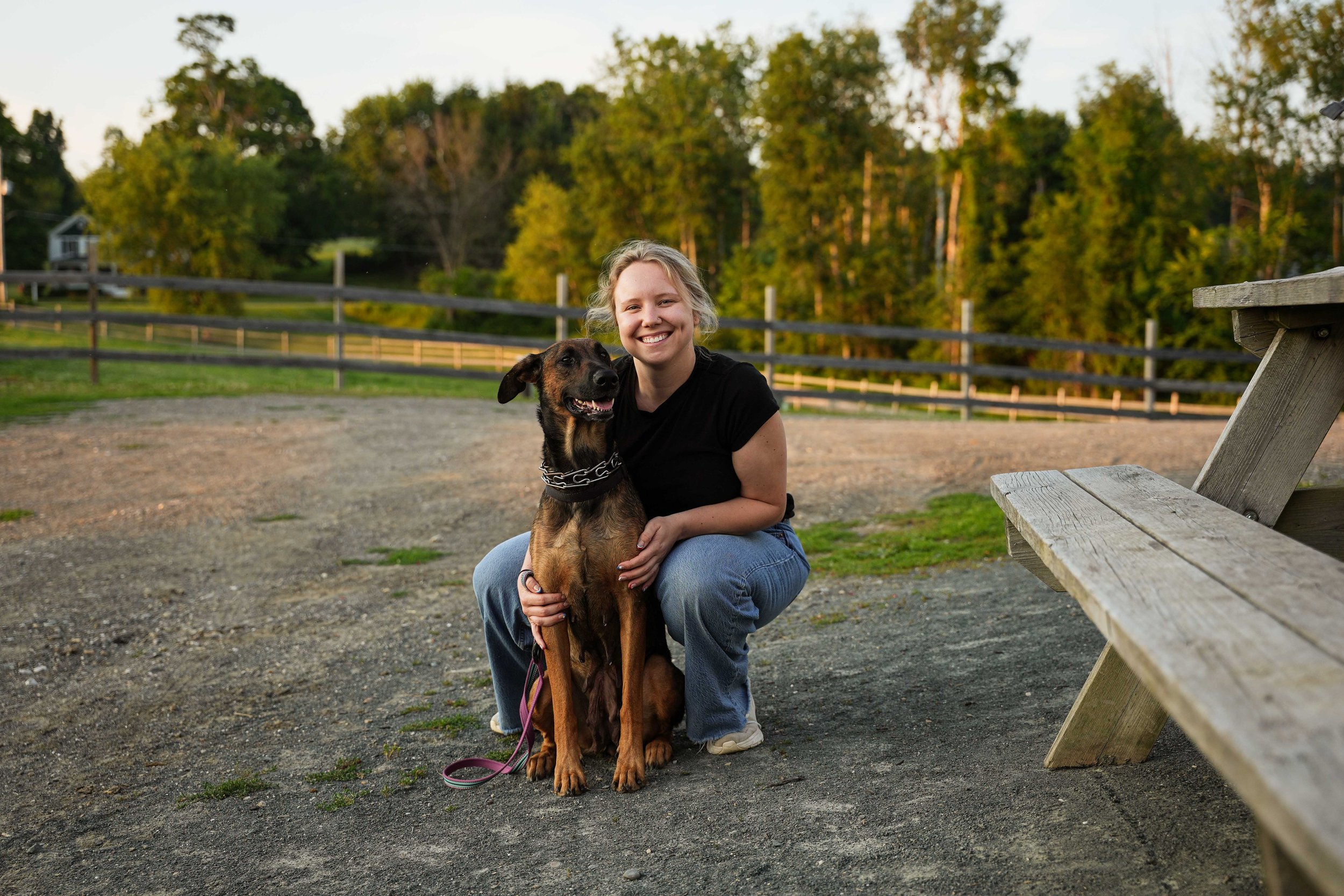 A young woman with blonde hair and a black shirt is kneeling on the ground, smiling, and hugging a brown and black dog with a chain collar. They are outdoors in a park or farm setting with a wooden fence, green trees, and a gravel area in the background. The photo is taken during the daytime with natural light.