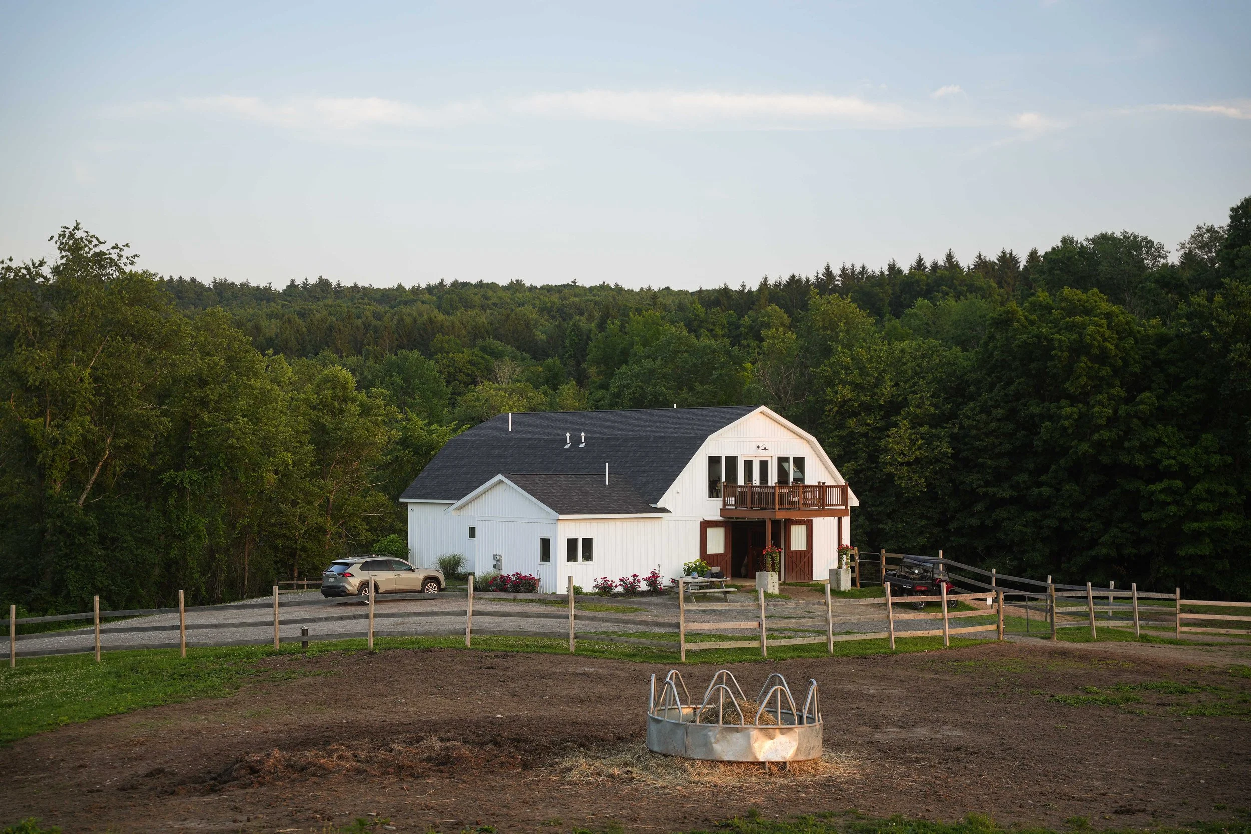 A white farmhouse with dark roof and a small balcony, surrounded by lush trees, with parked cars in front and a fenced garden area with a fire pit in the foreground.