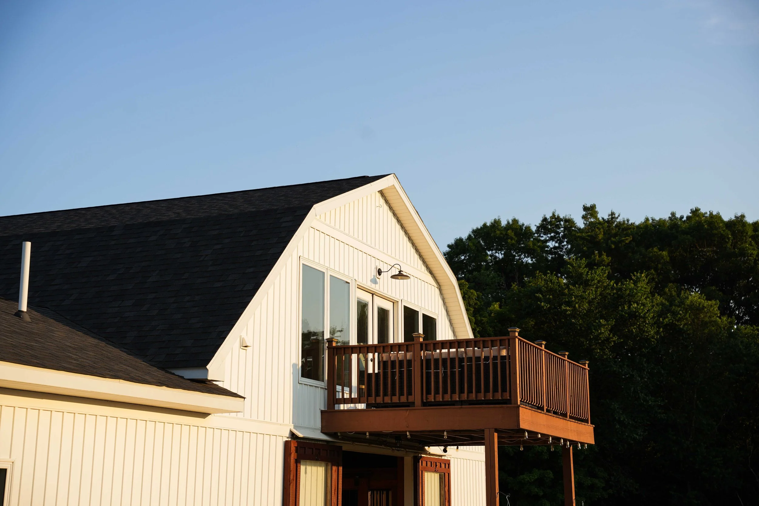 The image depicts a portion of a modern house with a sloped roof, large windows, and a wooden balcony or deck. There are trees in the background under a clear blue sky.