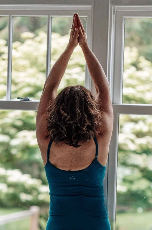 A woman practicing yoga, standing with her back to the camera, with her arms raised overhead and palms pressed together in front of a large window overlooking a green outdoor landscape.
