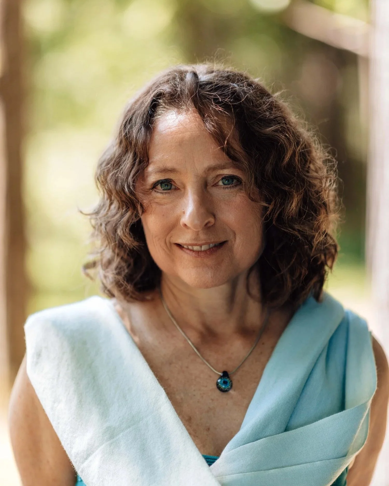 A woman with curly brown hair and blue eyes smiling outdoors, wearing a light blue outfit and a blue gemstone necklace.