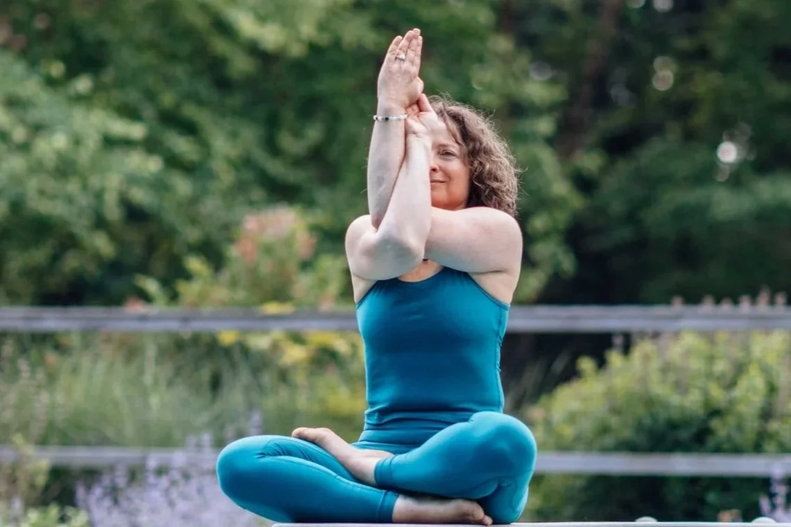 Woman sitting cross-legged outdoors practicing yoga or meditation with hands pressed together in a prayer position above her head, surrounded by greenery.