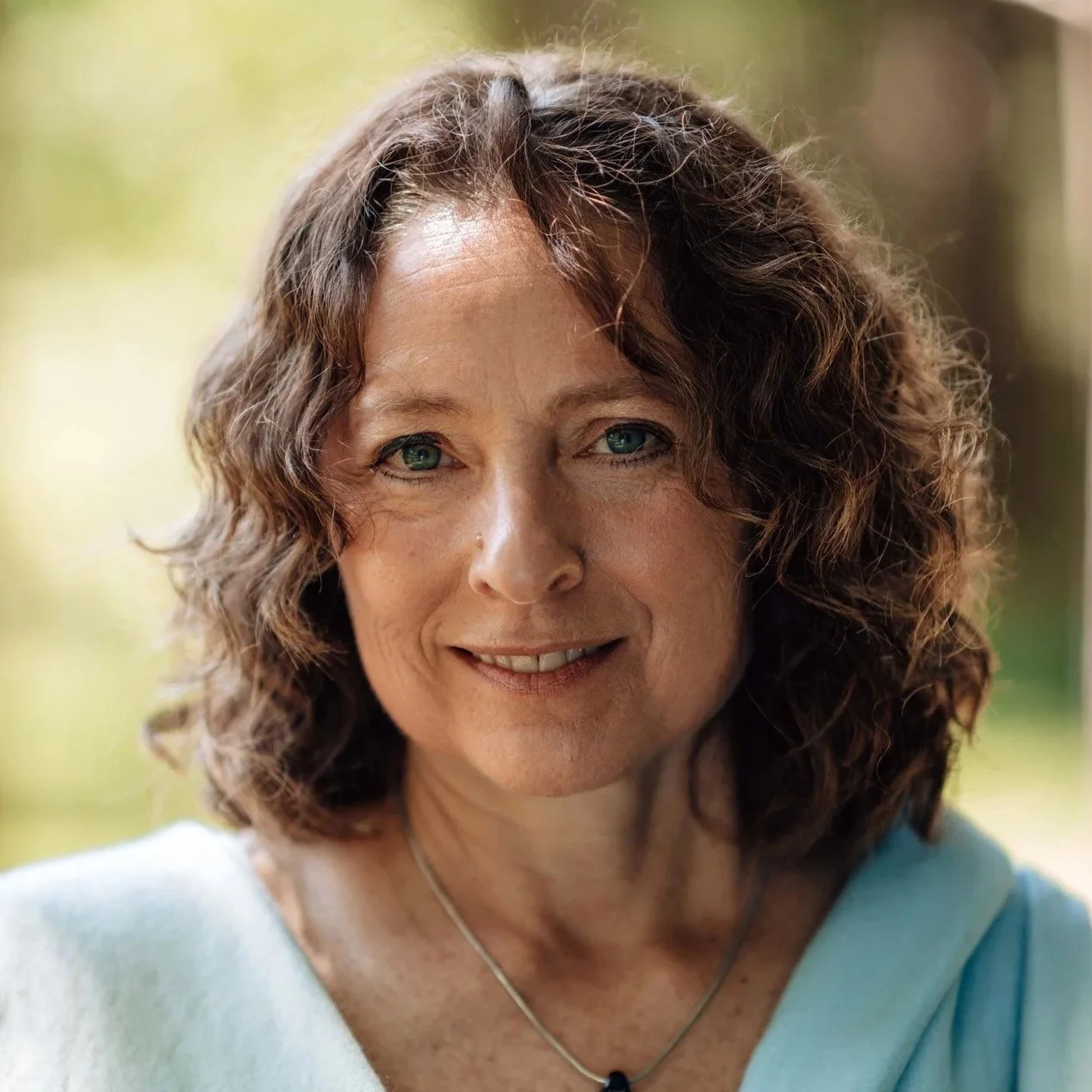Close-up of a middle-aged woman with curly brown hair and blue eyes, smiling, outdoors in natural lighting.
