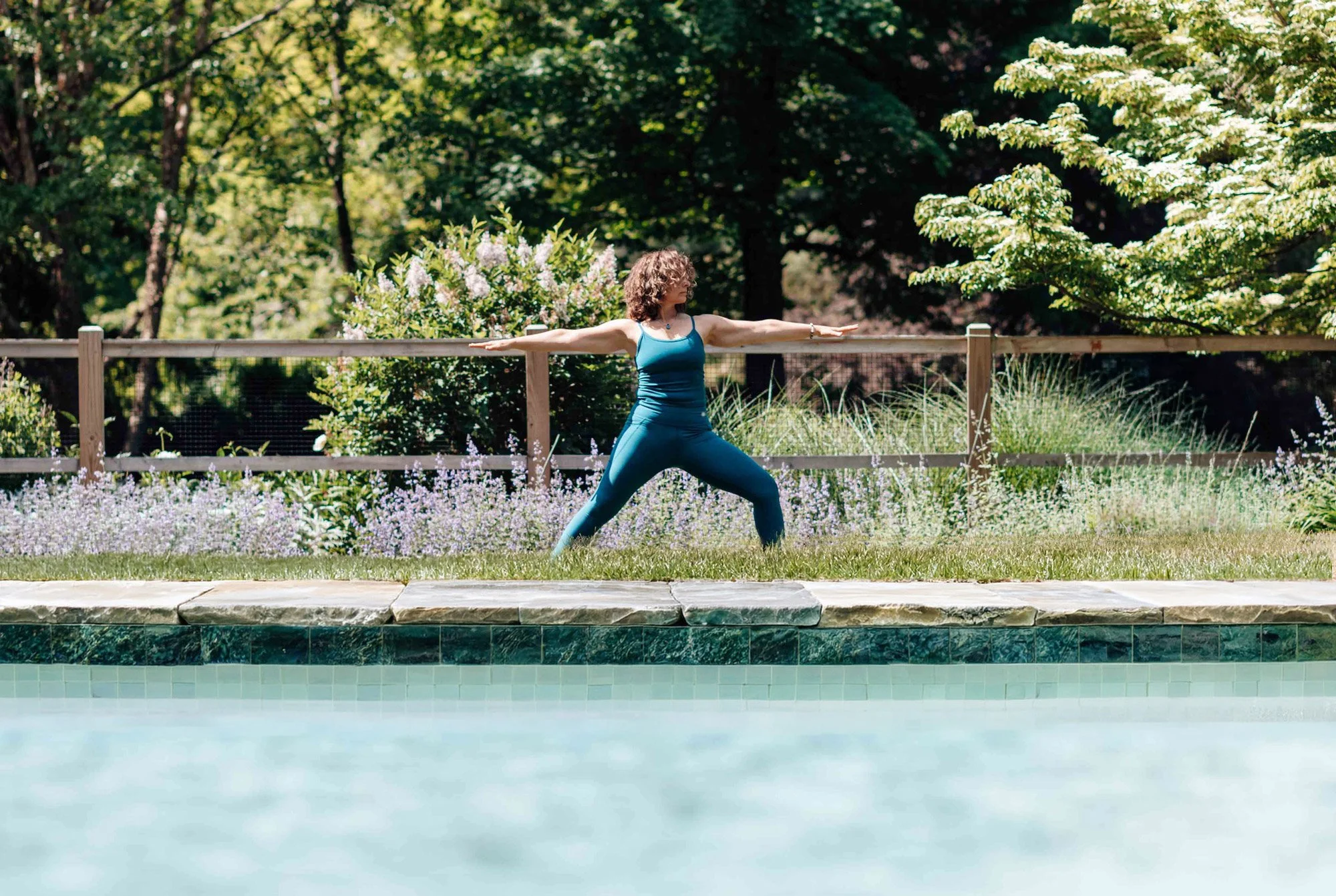 A woman practicing yoga outdoors in a garden with lush green trees and flowering bushes, standing on a stone path near a swimming pool.
