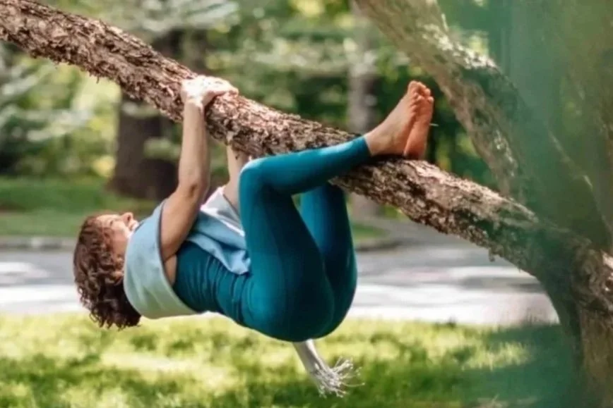 Person hanging upside down from a tree branch in a park or forest, holding onto the branch with both hands, wearing a blue sleeveless top and matching leggings.