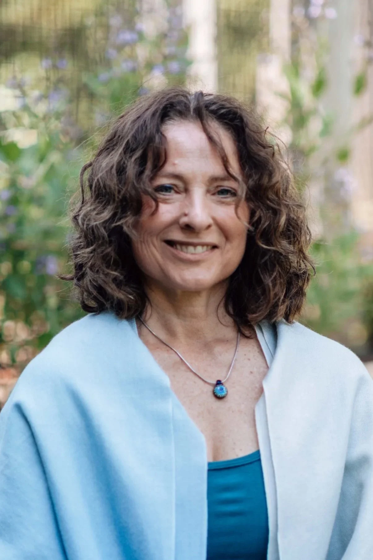 Smiling middle-aged woman with curly brown hair wearing a blue top and a matching turquoise necklace, outdoors with blurred greenery background.