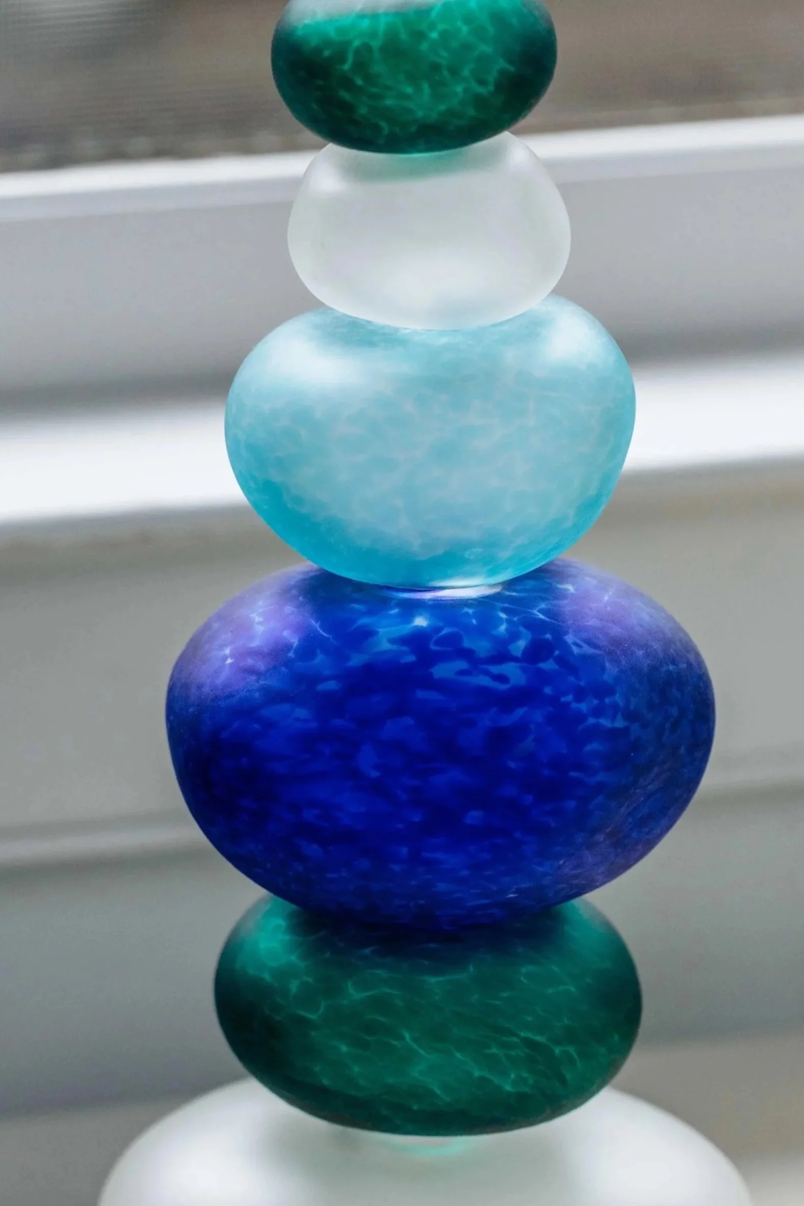 A stack of six smooth, rounded stones in various shades of blue and green, balanced on a white base, with a blurred background.