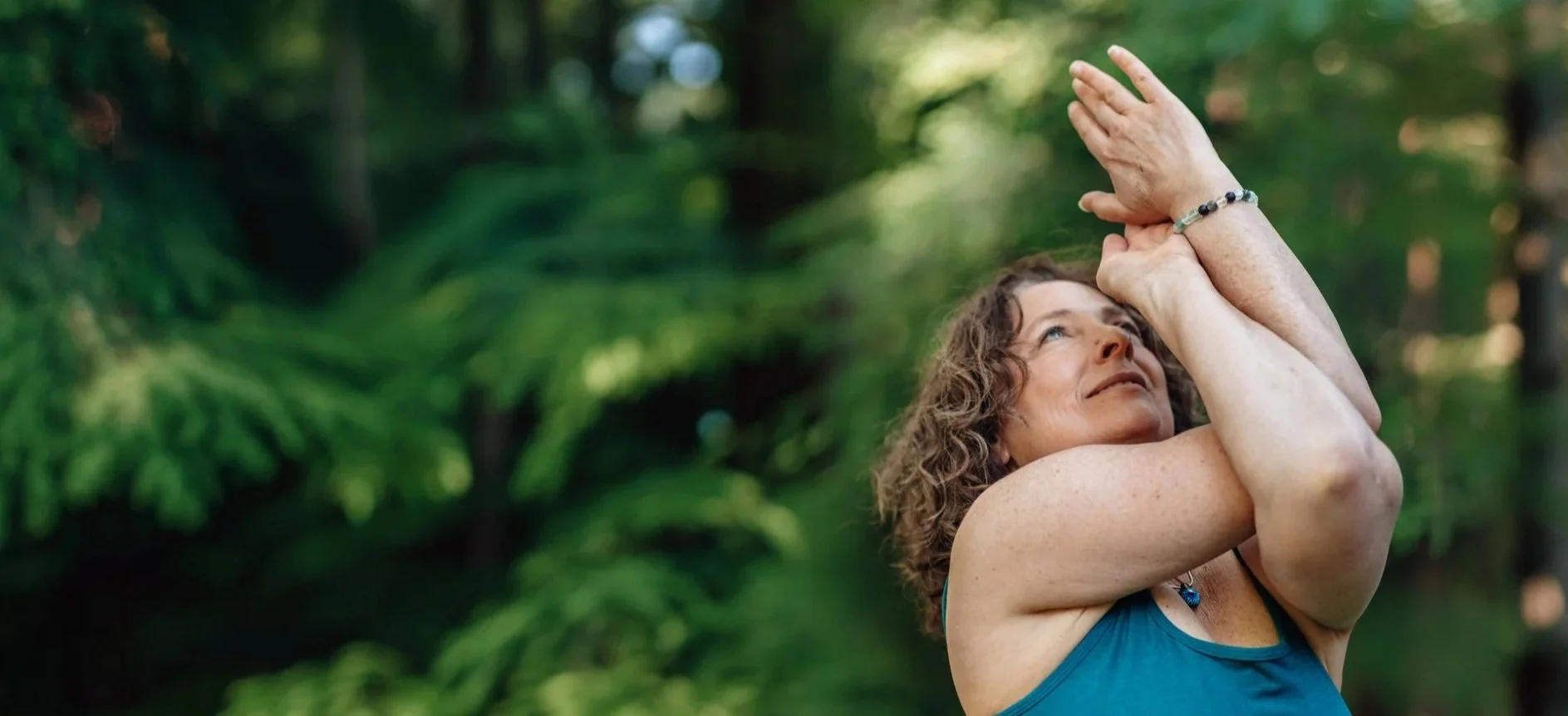 Woman with curly hair practicing yoga outdoors with arms raised and hands together in a prayer position, surrounded by lush green foliage.