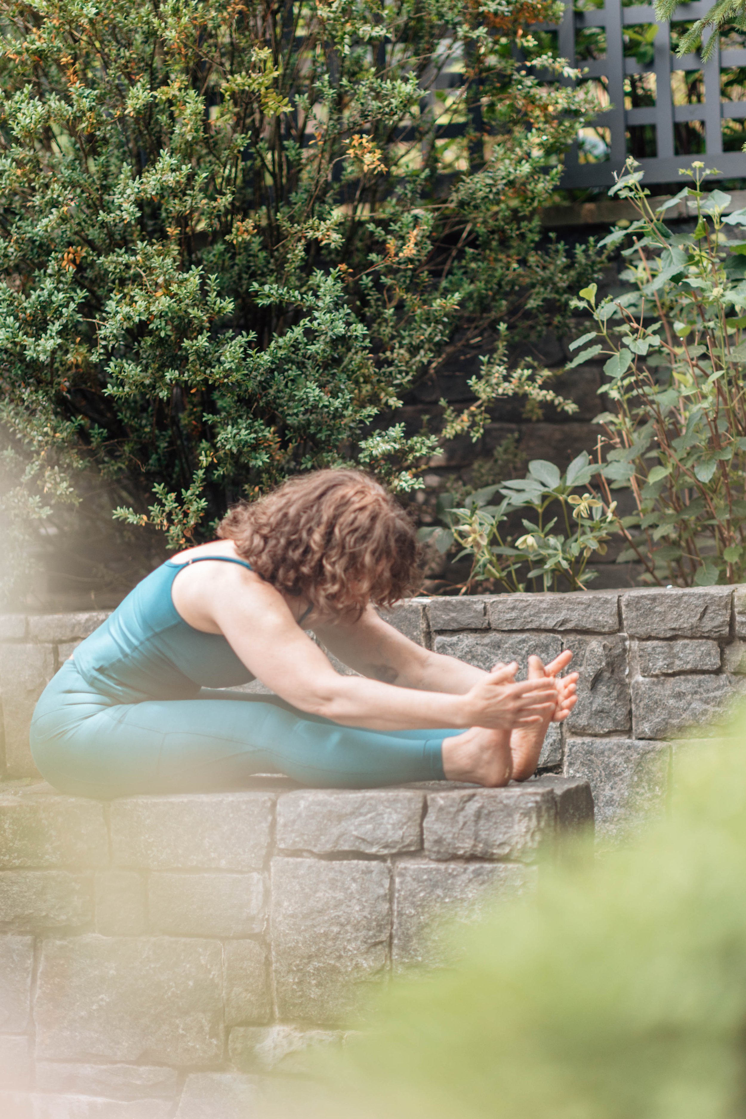 A woman practicing yoga outdoors on stepped stone platform, wearing a blue sleeveless top and matching leggings, surrounded by green leafy plants and a black lattice fence in the background.