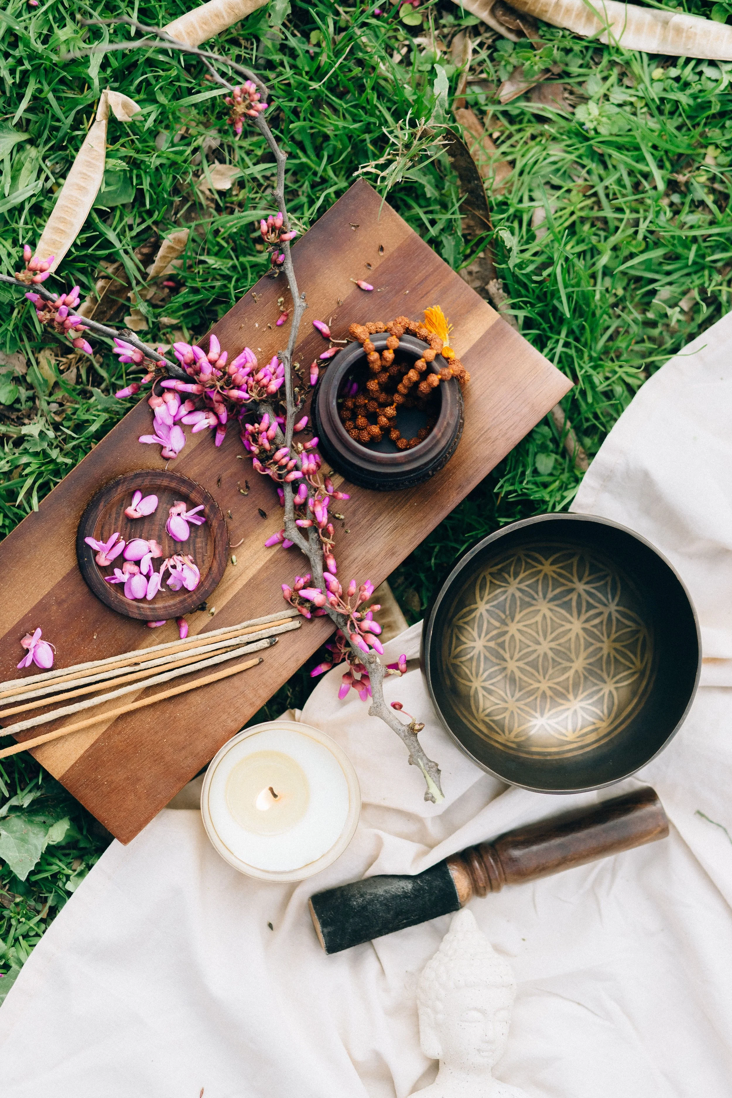 A flat lay of spiritual and meditation items on green grass, including pink flowering branches, candles, a black singing bowl, and a white Buddha head sculpture.