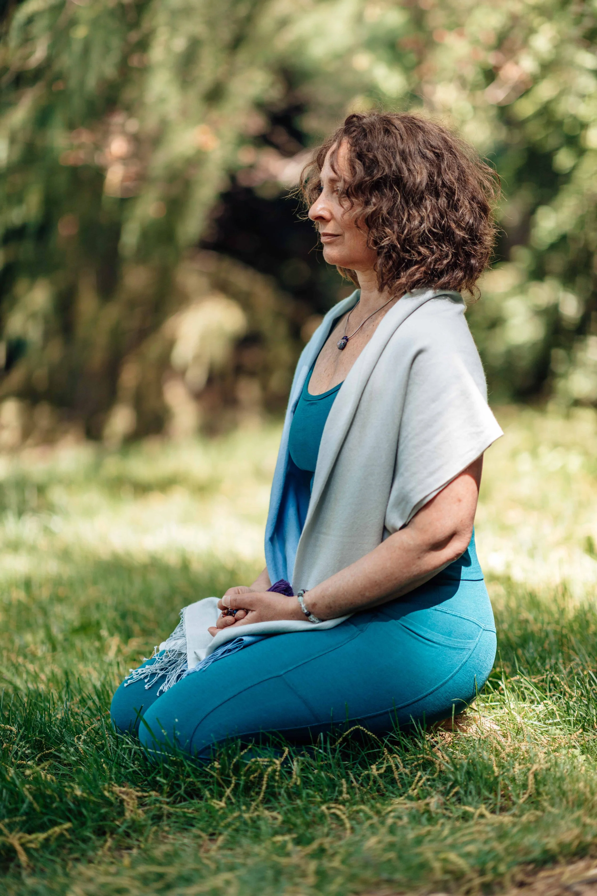 A woman kneeling in a grassy outdoor setting, meditating with her eyes closed, wearing a teal shirt, light gray vest, and blue jeans, surrounded by trees with sunlight filtering through.