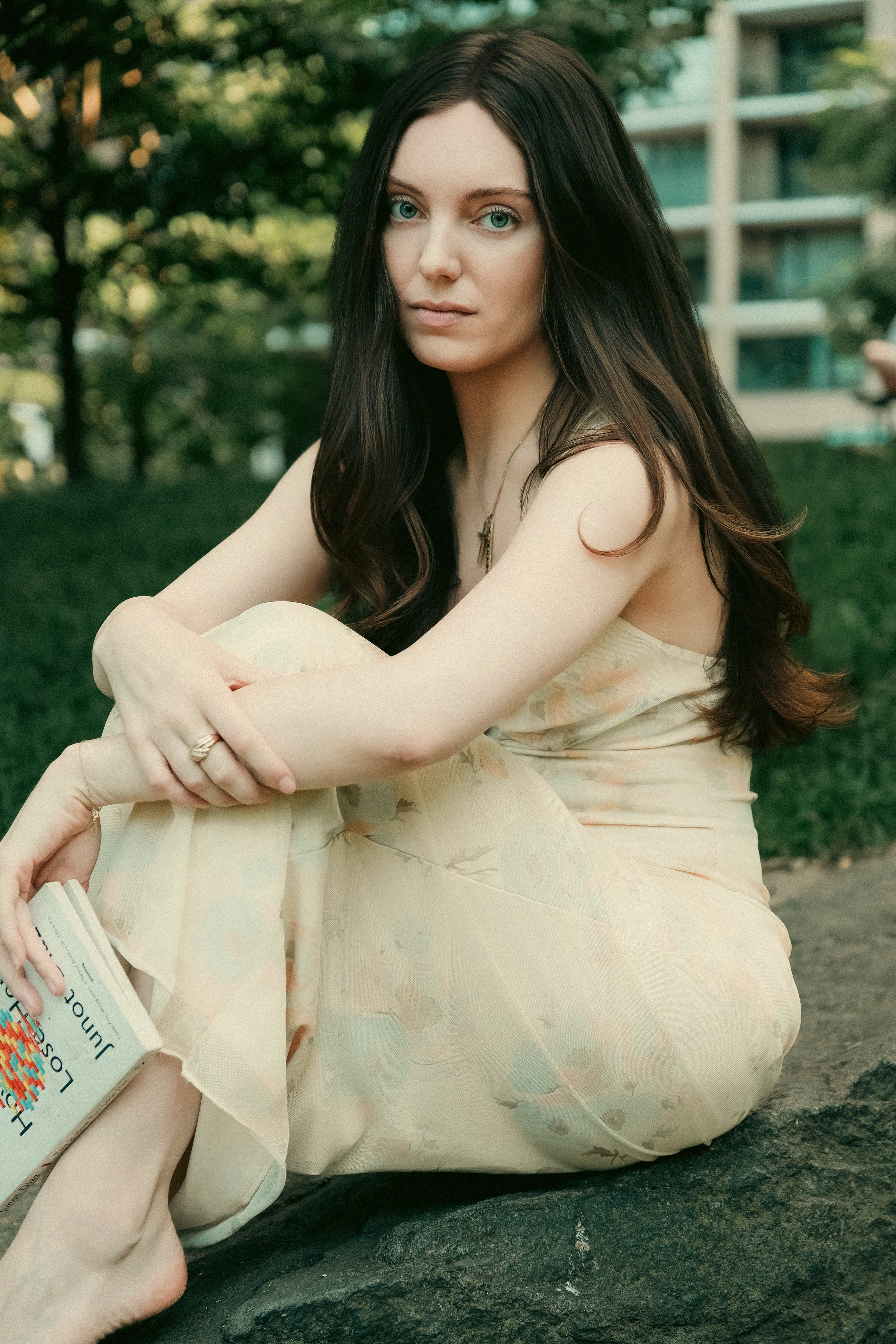 A young woman with long dark hair and light skin sitting outdoors surrounded by green trees and a building in the background. She is wearing a light floral dress and looking directly at the camera with a neutral expression, holding a book in her lap.