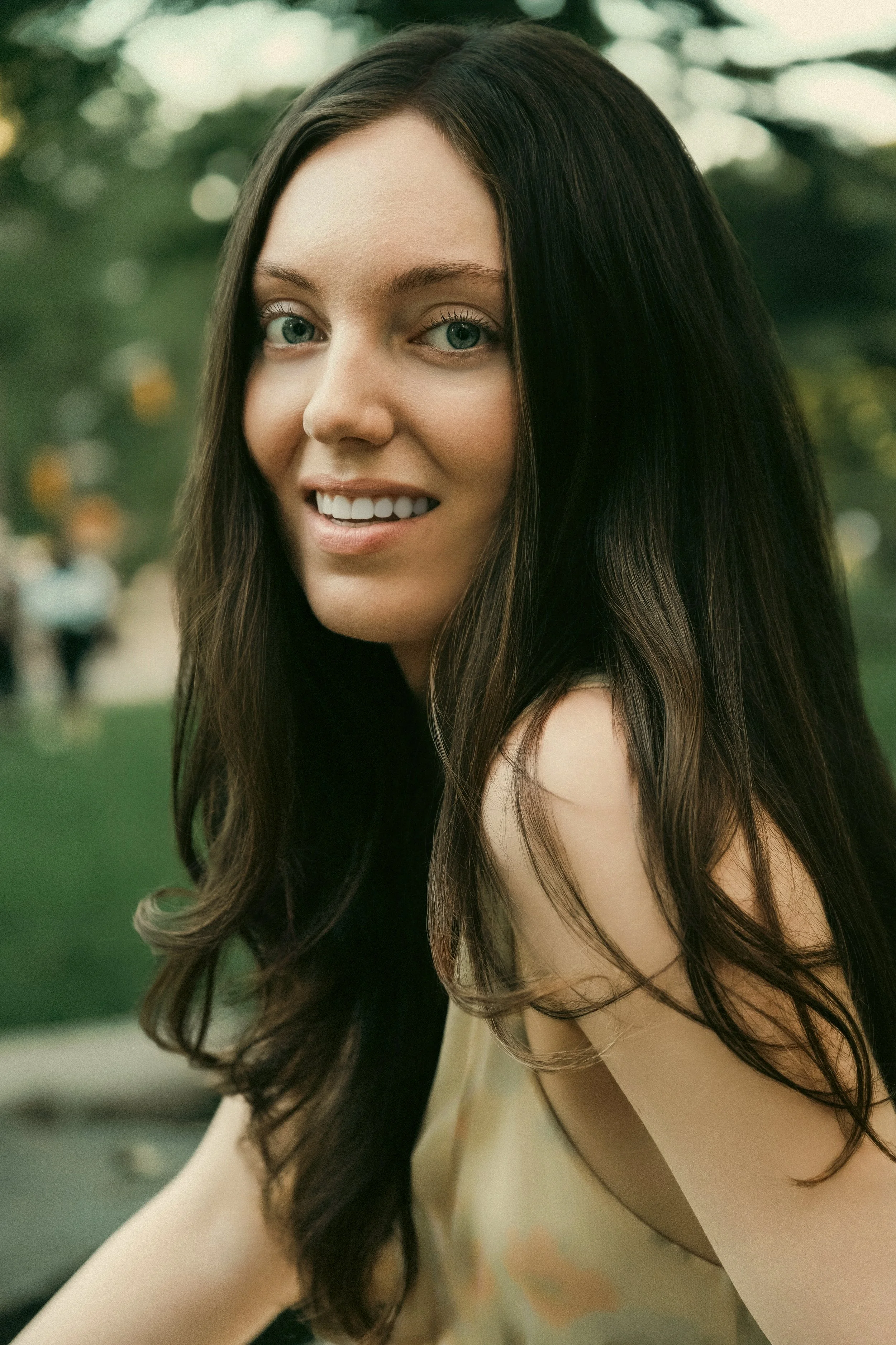 A young woman with long brown hair and blue eyes smiling outdoors, blurred greenery in the background.