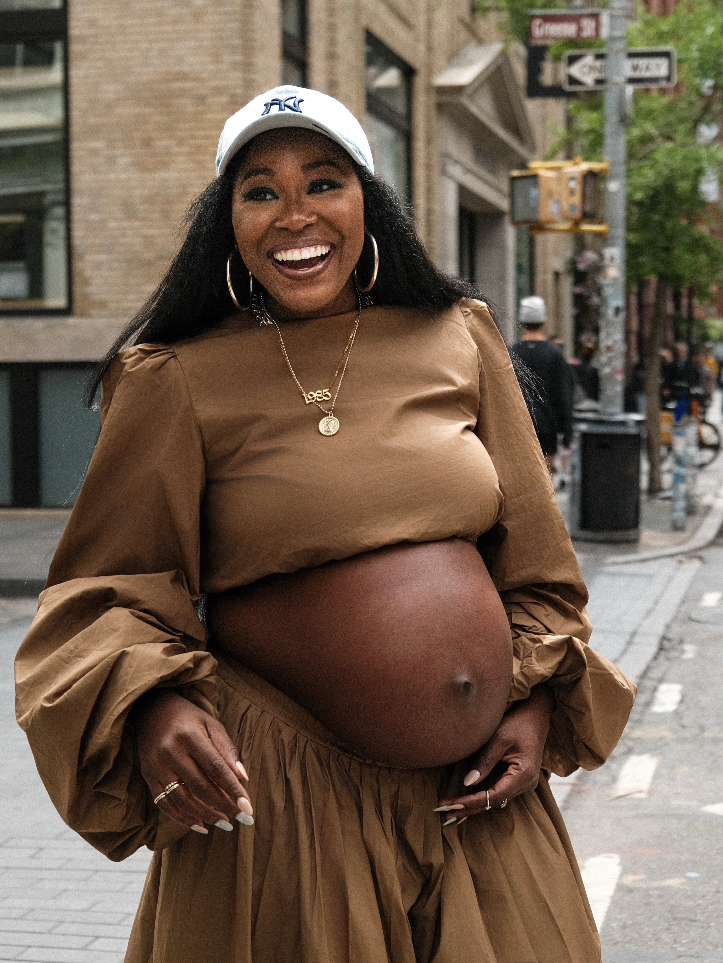 A pregnant woman wearing a white baseball cap, hoop earrings, and layered necklaces, standing outdoors on a city street, smiling broadly with her hand resting on her belly.