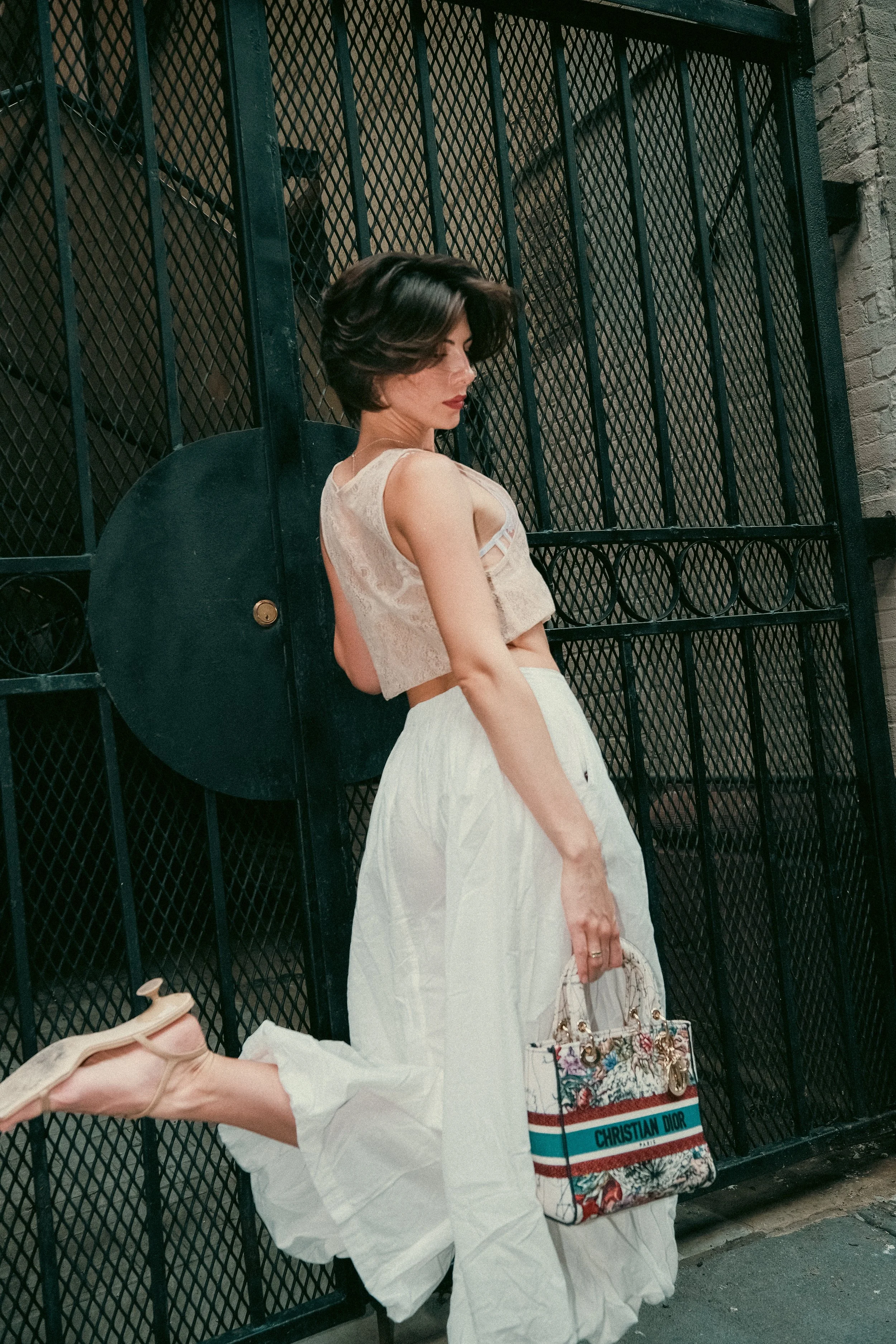 A woman with dark hair and a beige sleeveless top, standing in front of a black metal gate, holding a Christian Dior tote bag with colorful artwork, wearing white wide-leg pants and beige shoes.