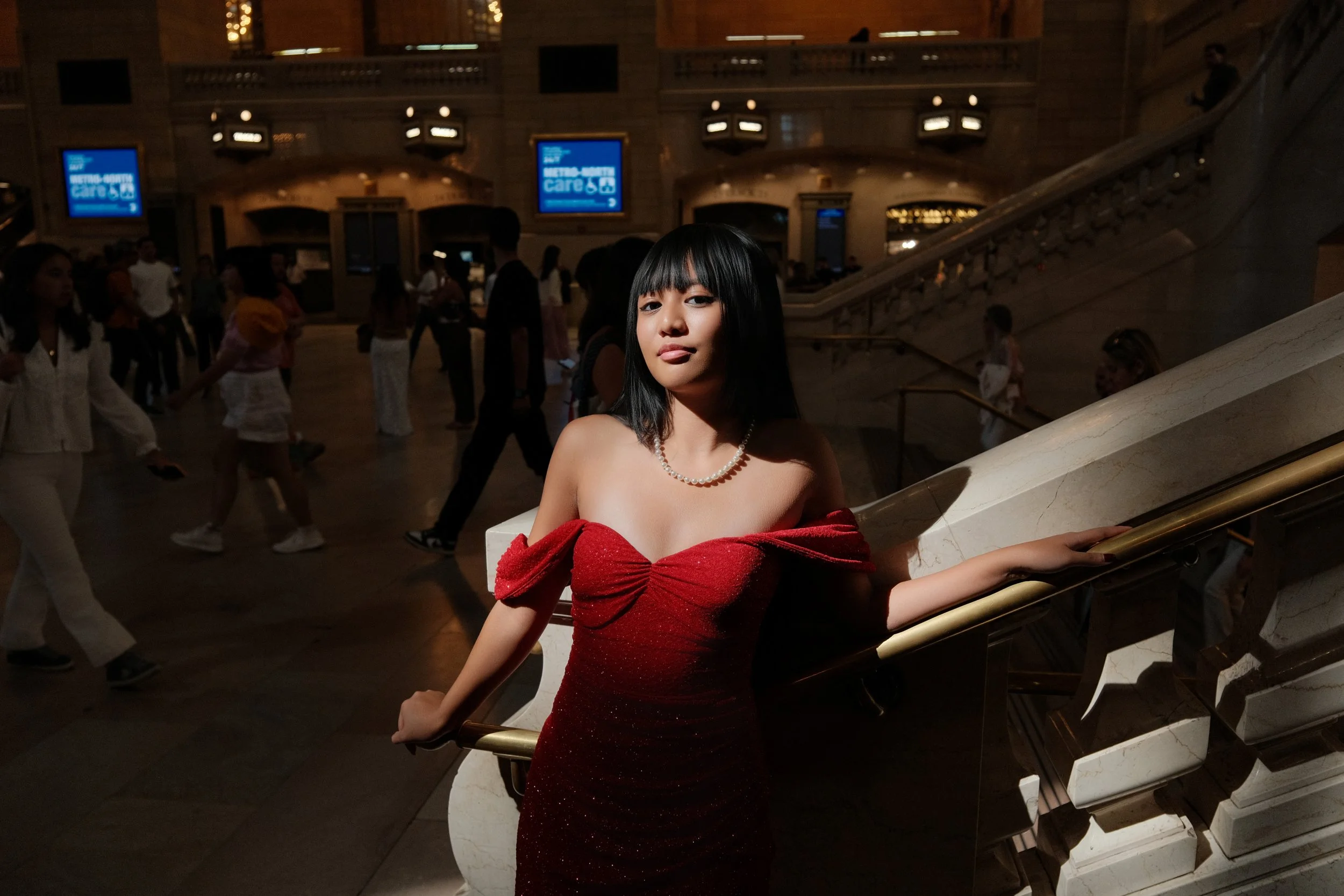 A woman in a red dress standing on a staircase inside a busy train station or airport terminal.