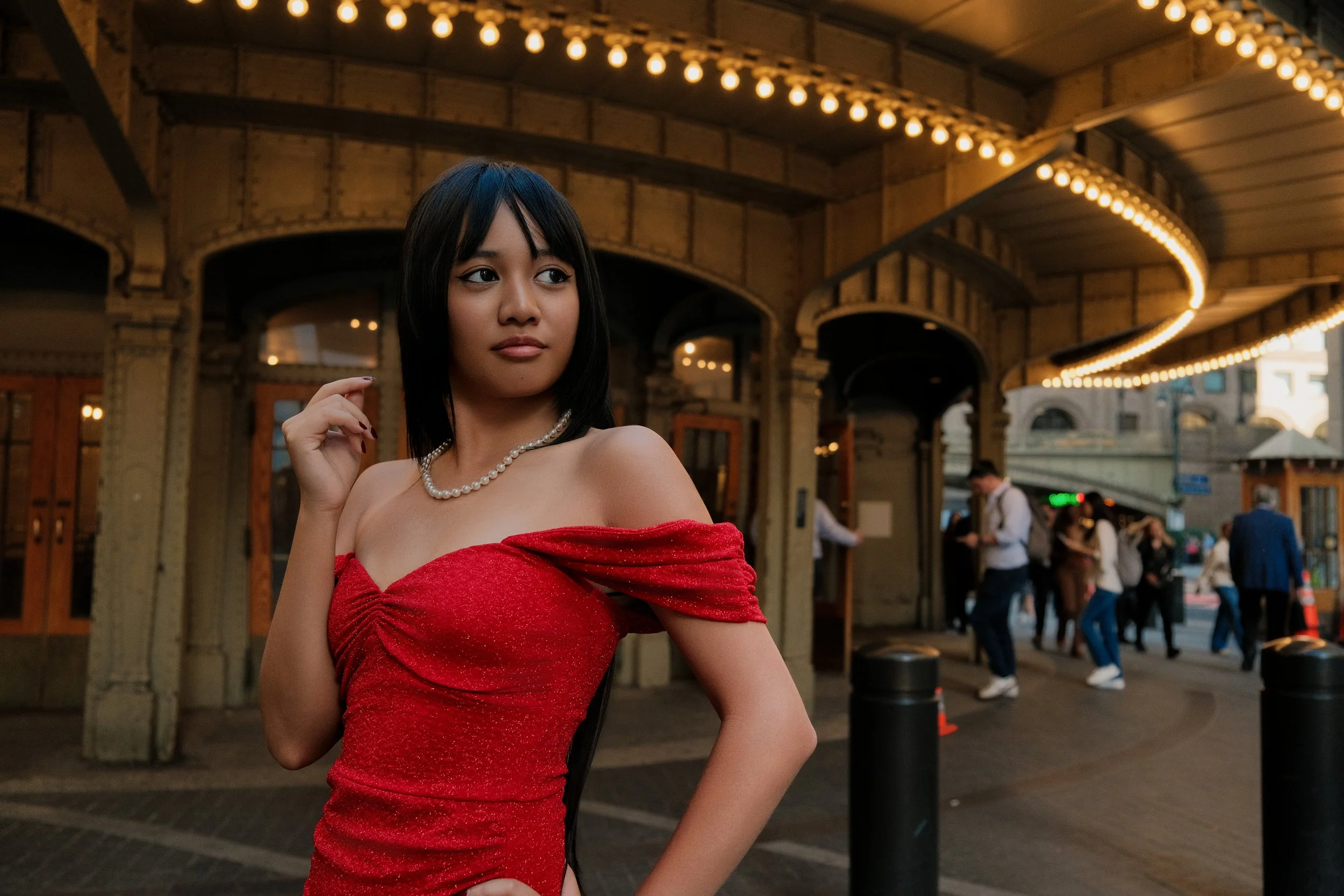 A woman with dark hair wearing a strapless red dress and pearl necklace standing under string lights in an outdoor setting with a crowd in the background.