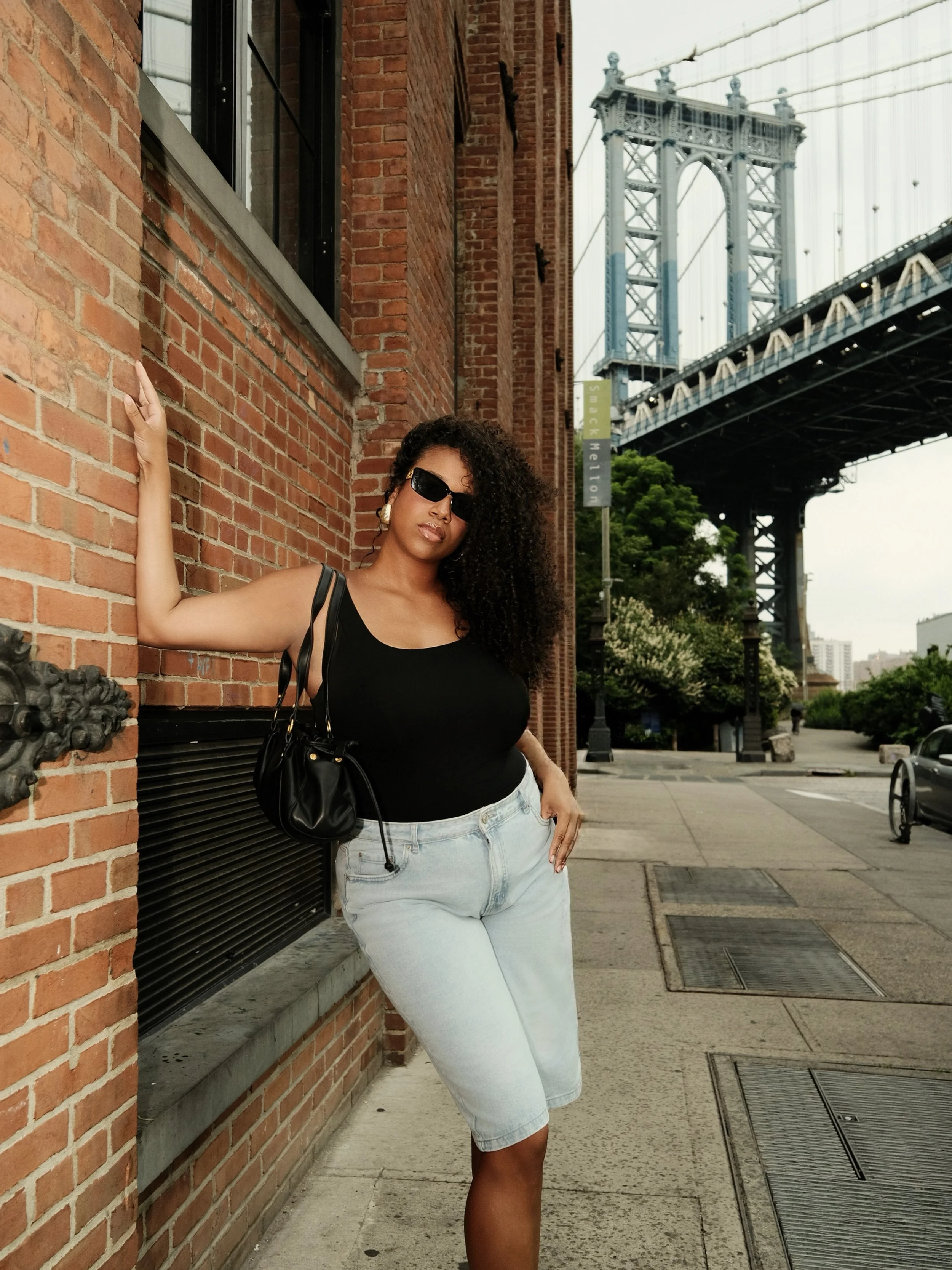 A woman with curly hair, sunglasses, black top, and light jeans, leaning against a brick building with a bridge in the background.