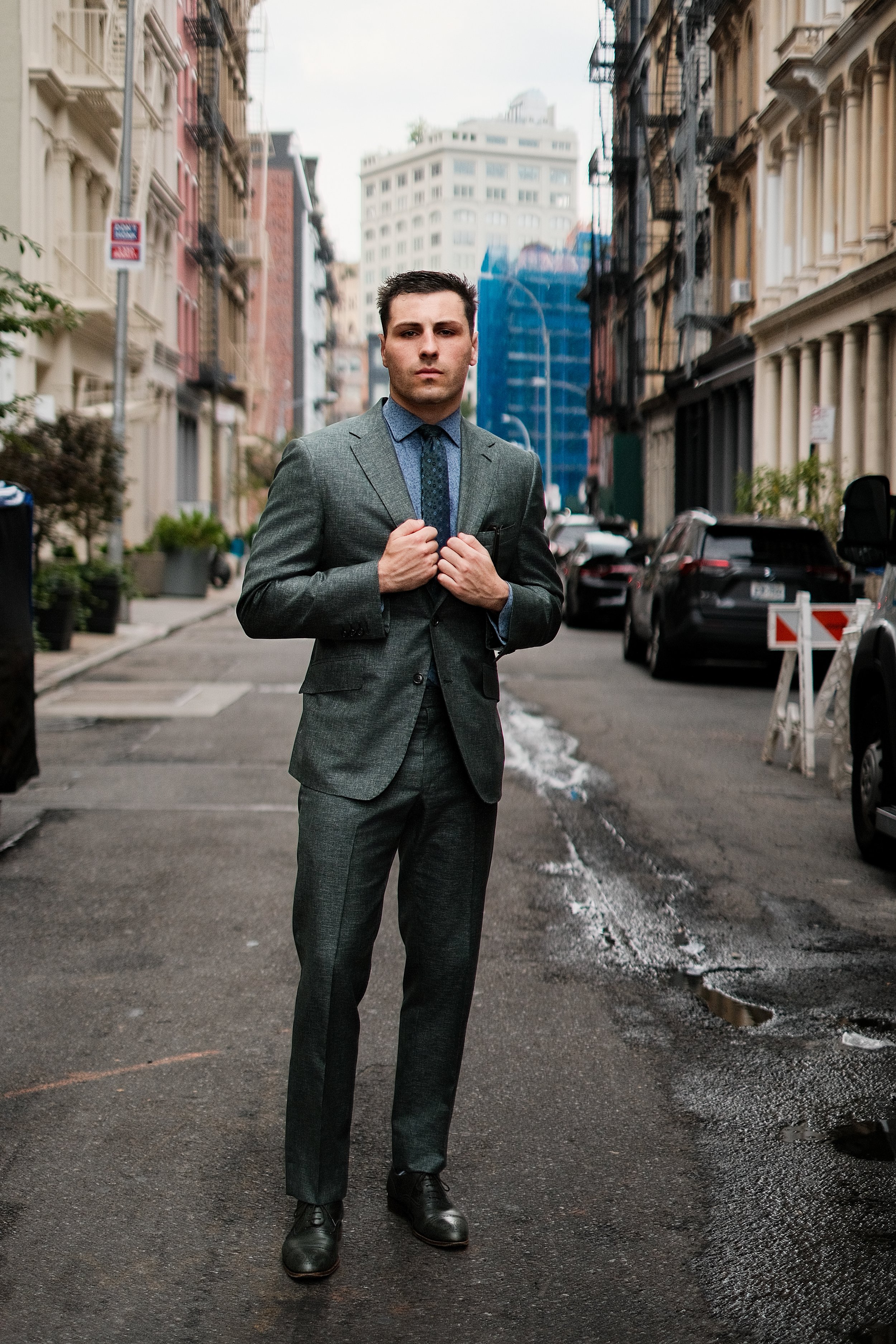 A young man in a gray suit and blue shirt standing in the middle of a city street, adjusting his jacket. Tall buildings line both sides of the street, and there are parked cars along the curb. The street appears damp with patches of snow or ice on th