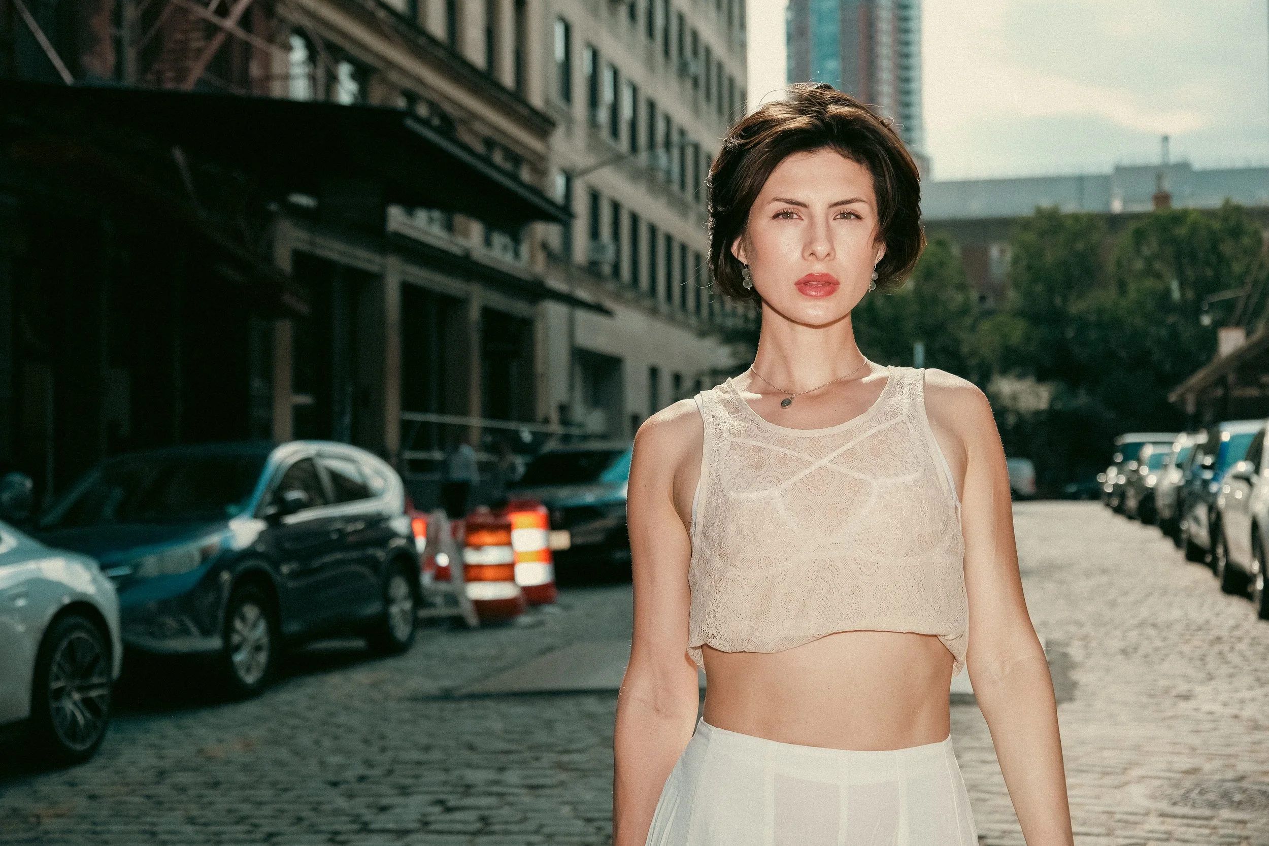 A woman with short dark hair and fair skin standing on a city street with parked cars and buildings in the background, wearing a sleeveless lace top and a necklace.