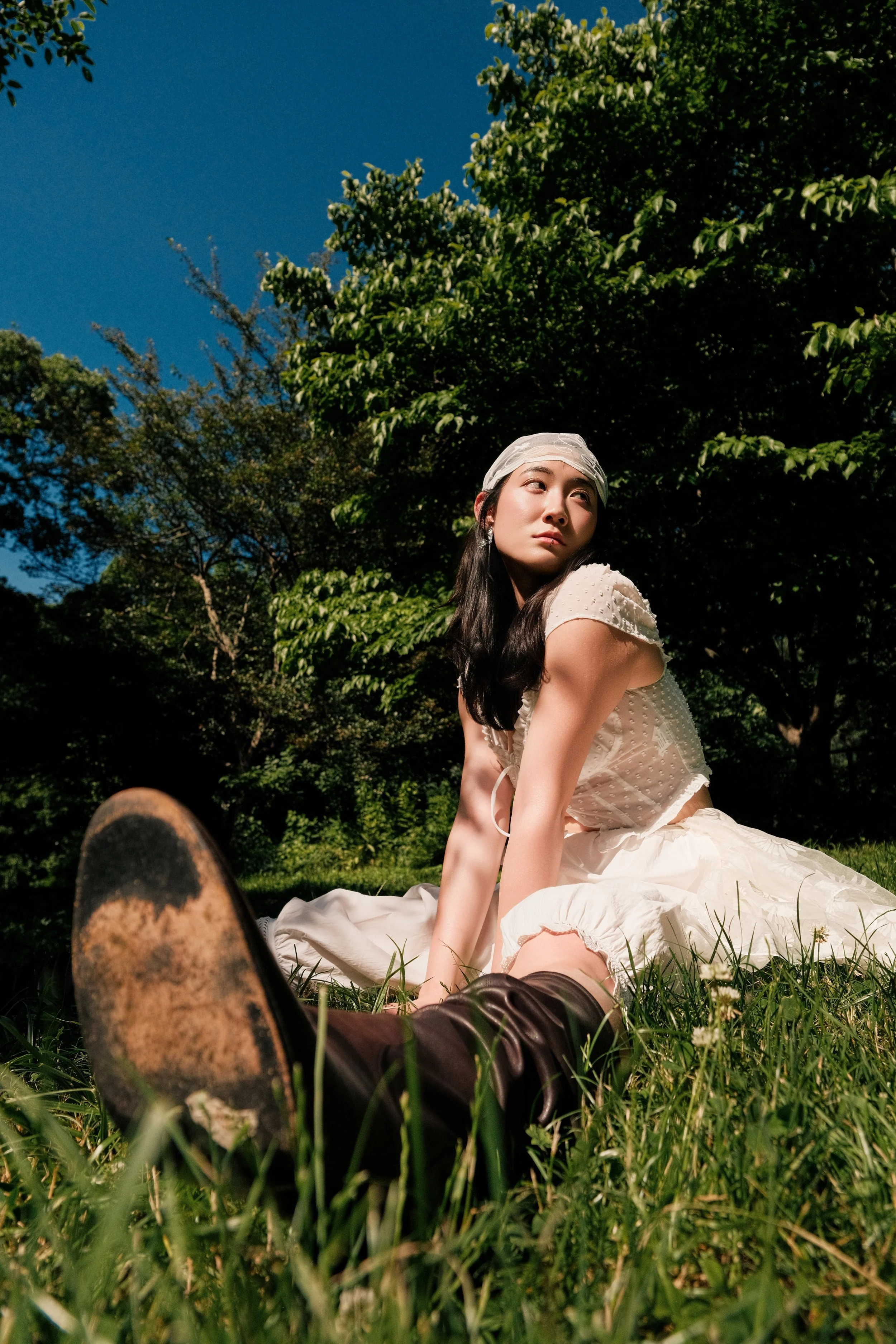 A woman sitting on the grass outdoors, looking to the side with a neutral expression. She is dressed in a light-colored dress and has a white headscarf. Trees and a clear blue sky are in the background.
