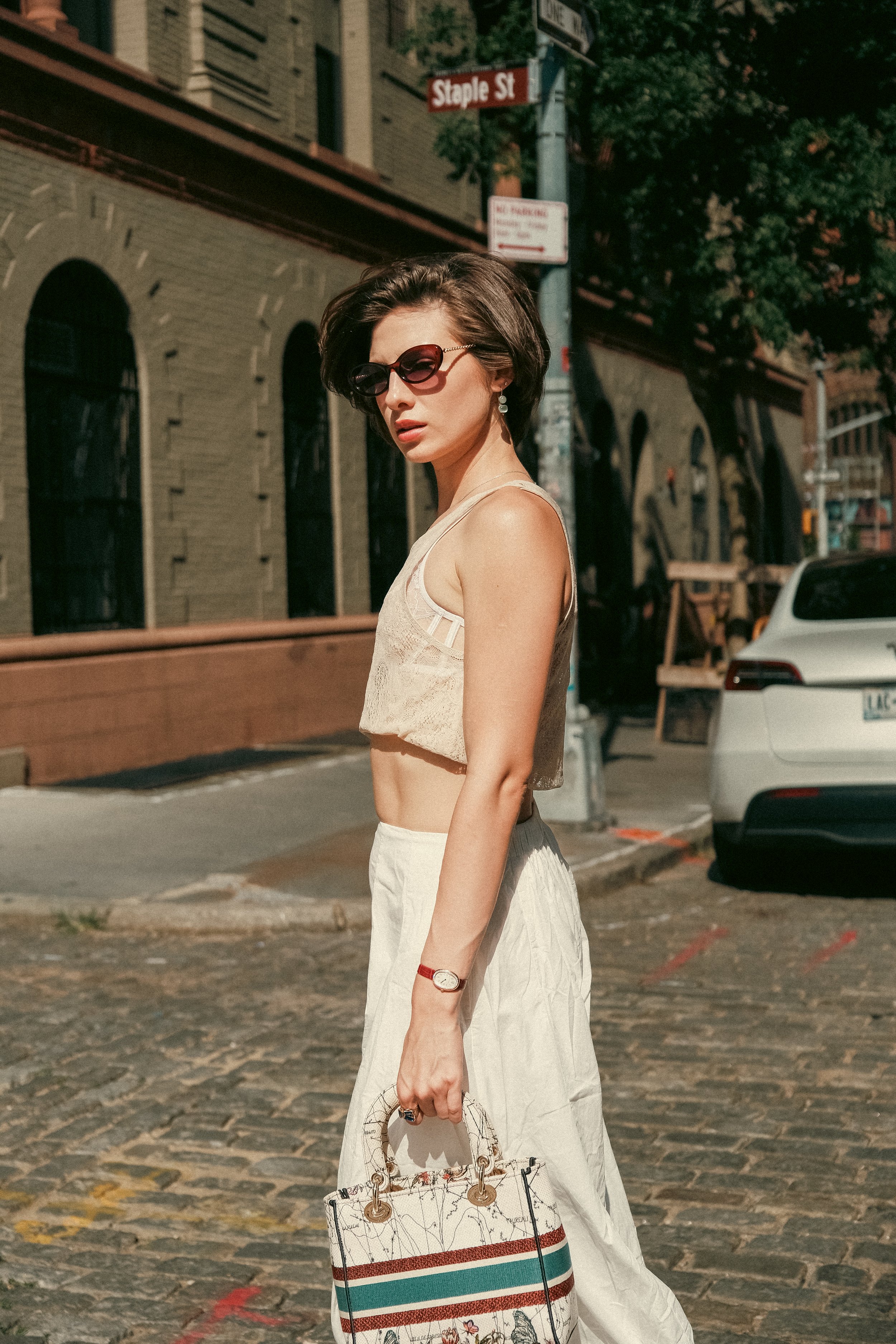 A woman wearing sunglasses, a cream sleeveless top, a white skirt, and a red watch is standing on a city street, holding a patterned handbag. The background features brick buildings, parked cars, and street signs, including a sign that says 'Staple S