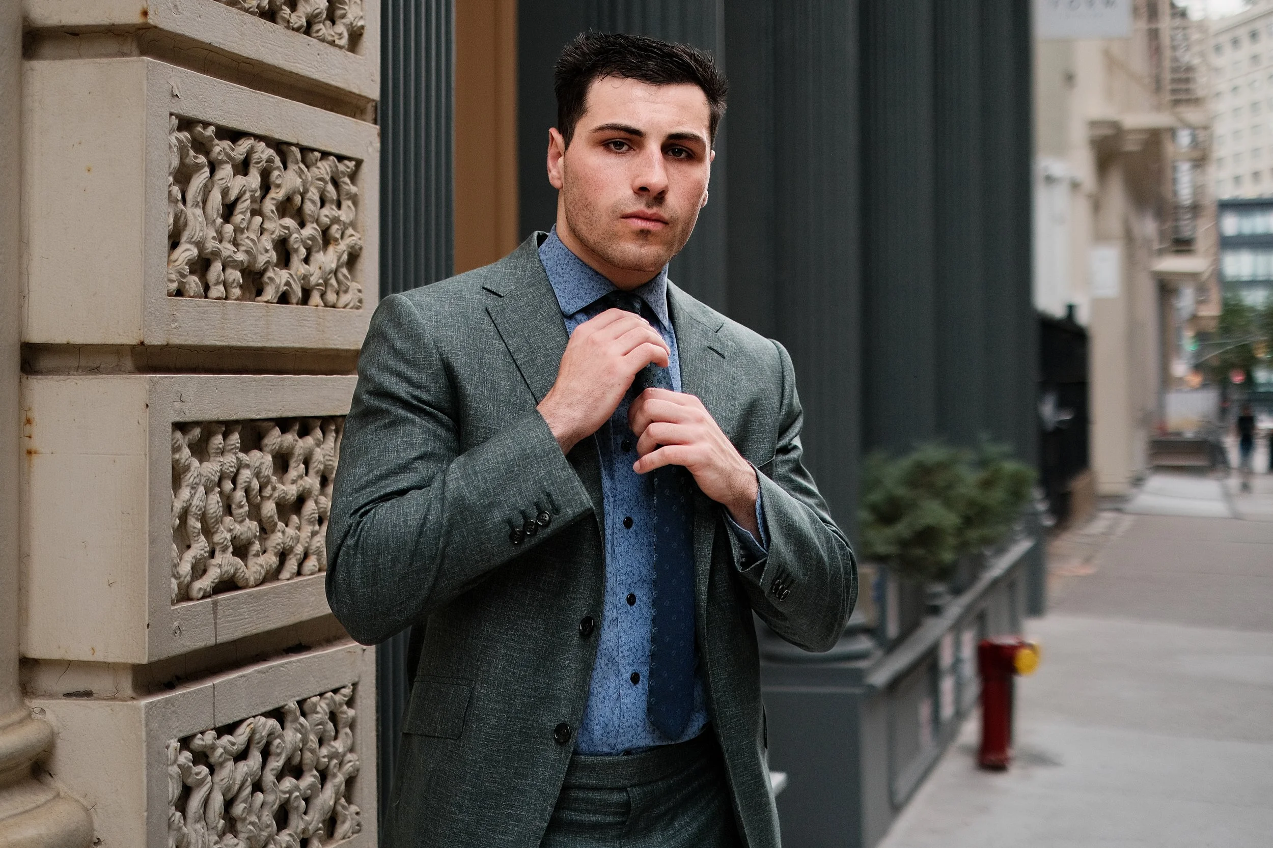 A man in a gray suit adjusting his tie on a city street with buildings in the background.