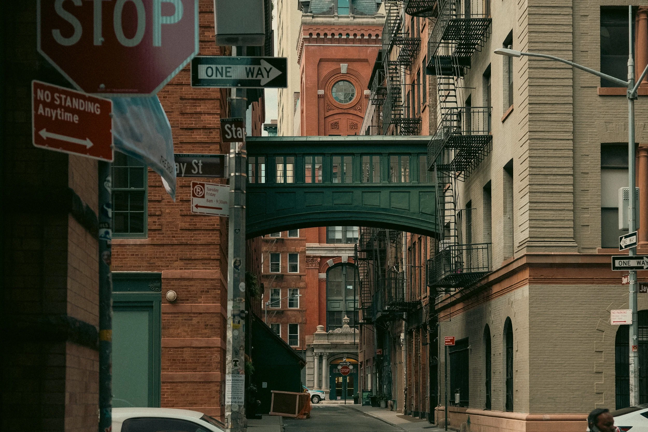 View of a city alleyway with brick and beige buildings, fire escapes attached to the buildings, and a green covered bridge or walkway connecting the buildings. Several street signs are visible, including a stop sign, one-way sign, and no standing sig