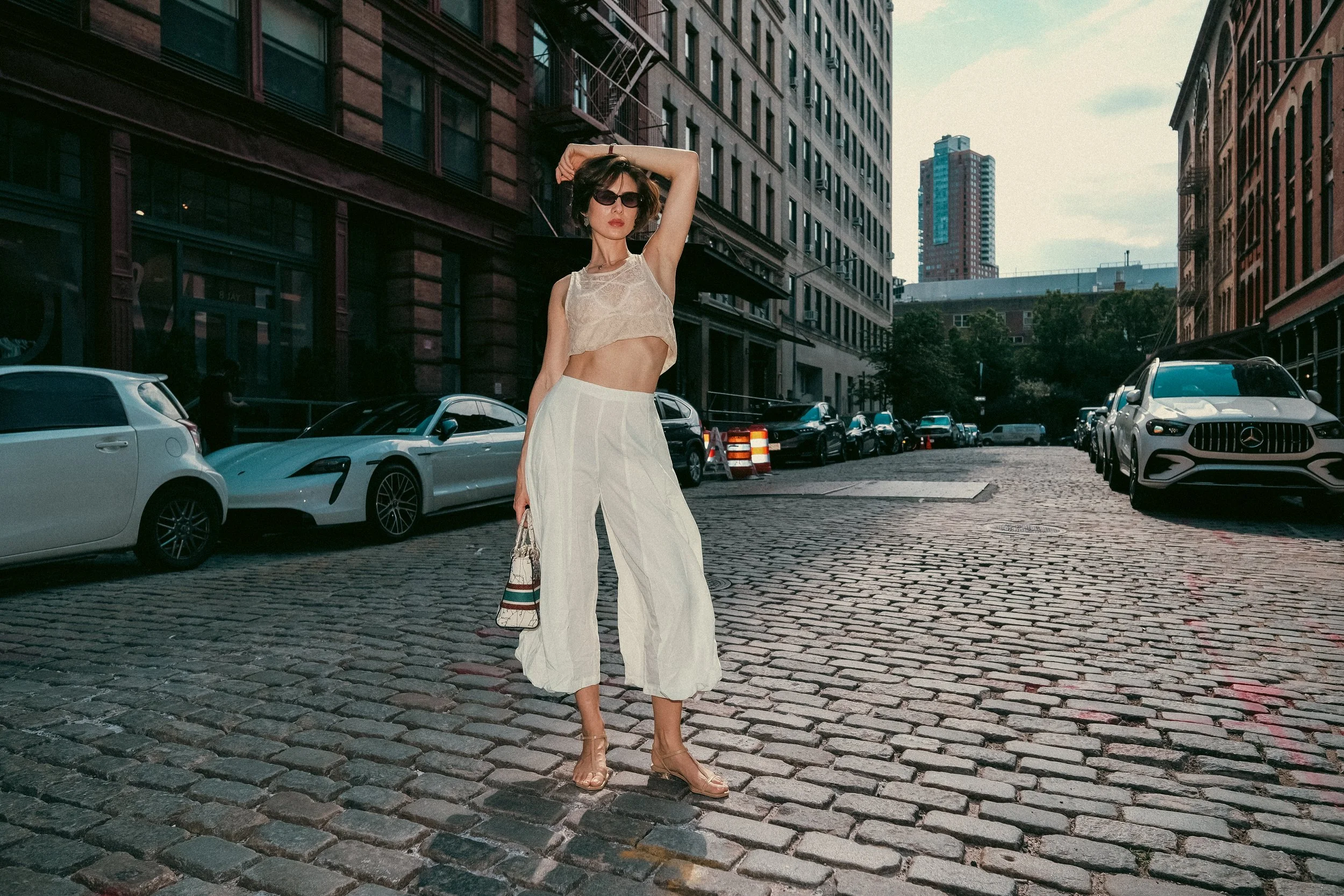 A woman with short dark hair, wearing sunglasses, a beige crop top, white wide-legged pants, and high heels, stands on a cobblestone city street lined with cars and tall buildings in the background.