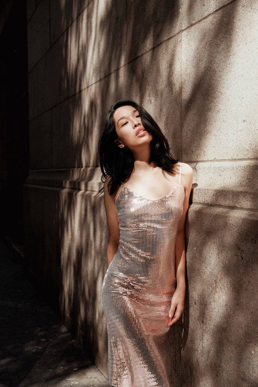 A woman with black hair wearing a shiny, pink, sequined dress leaning against a textured stone wall with shadows cast from nearby trees.