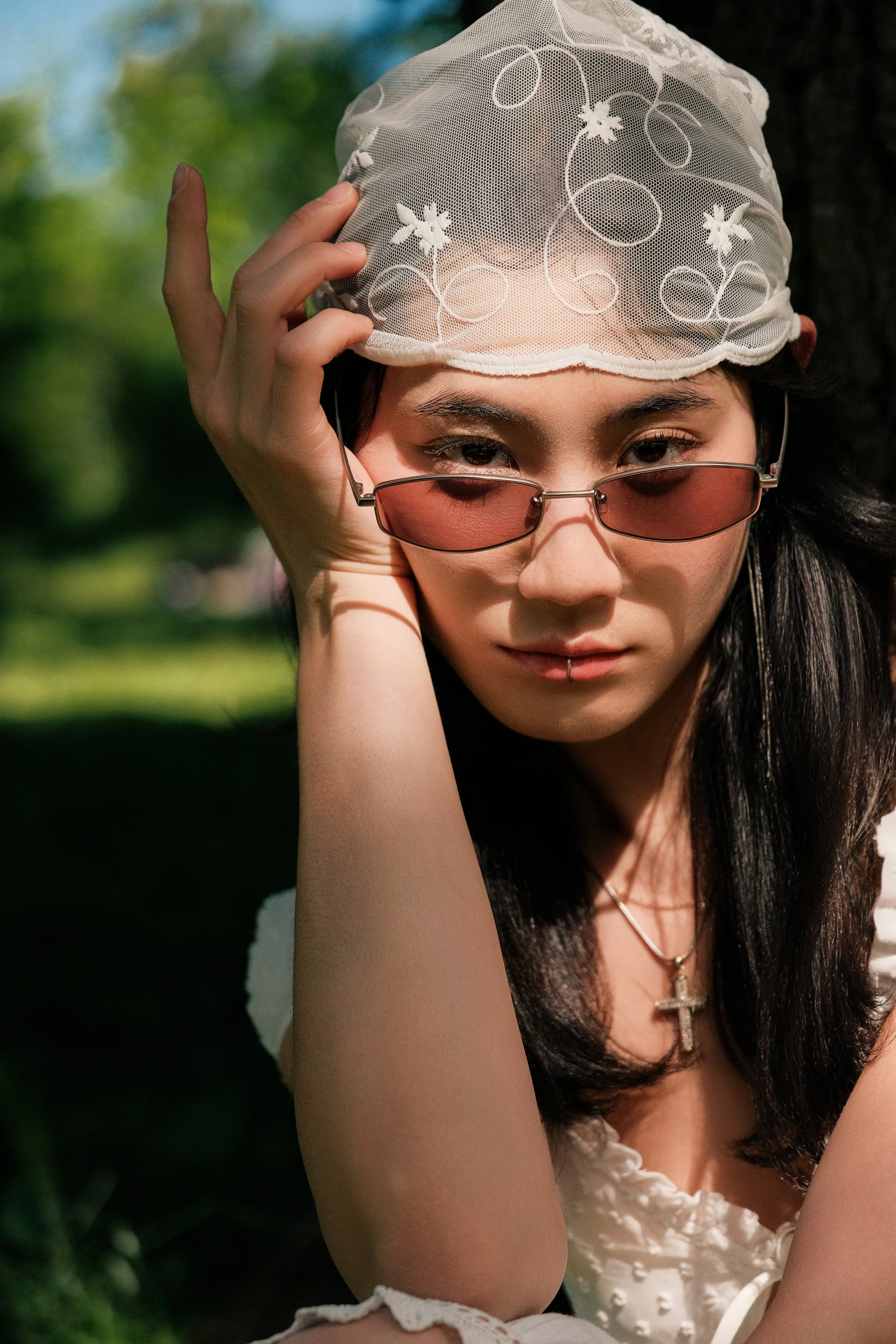 A woman wearing a white lace hat and pink-tinted sunglasses, with long dark hair, outdoors against a blurred natural background of greenery.